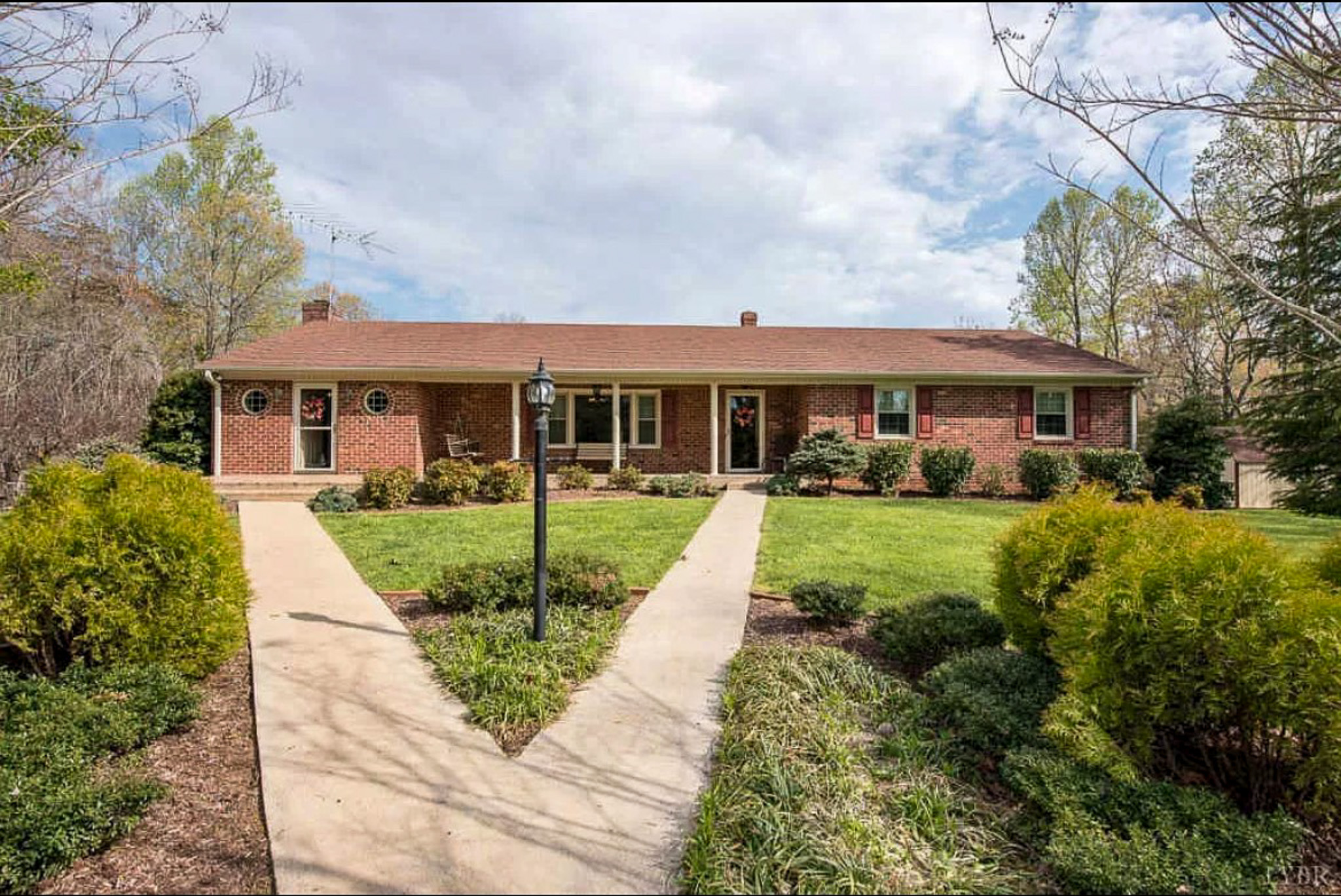 A brick ranch‑style house with a red roof sits behind a landscaped yard, with a V‑shaped walkway leading to a central lamppost and the front porch belonging to Sponsored Residential Providers Carolyn and Phil Ware in Amherst, Virginia.