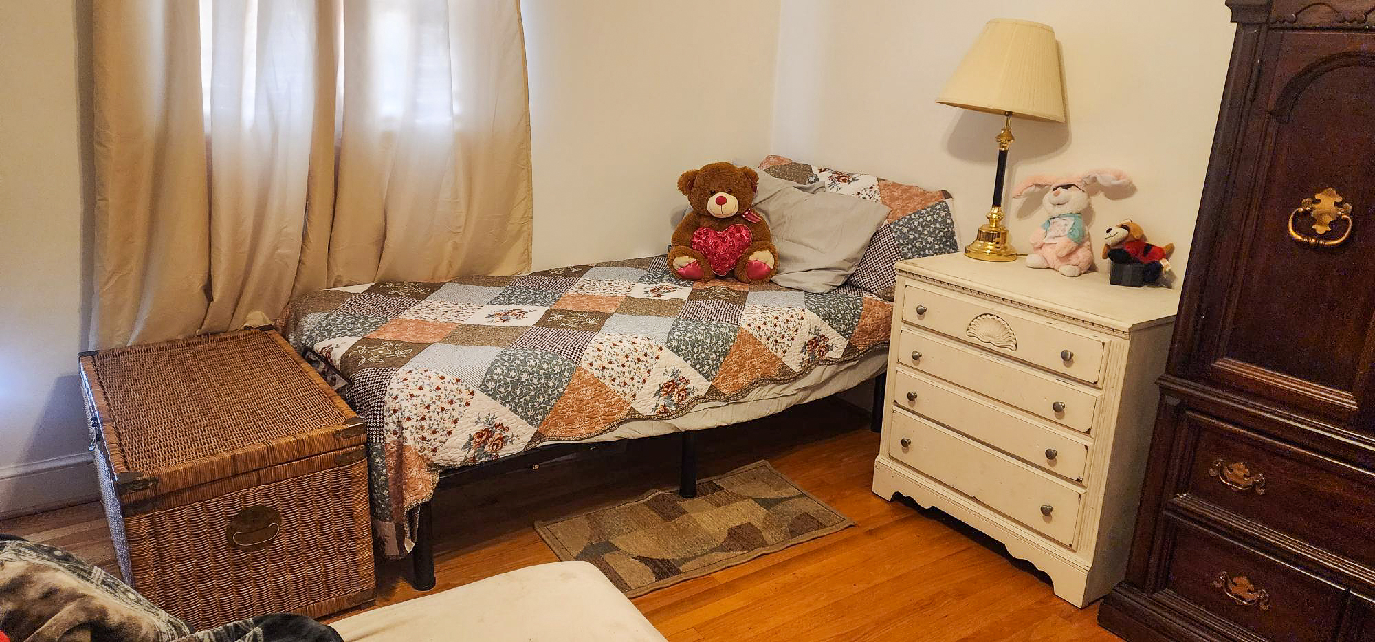 Small bedroom with a single bed topped with a patchwork quilt, stuffed animals on the pillows, a white dresser with a lamp, and wood floors inside the home of Sponsored Residential Provider Cindy Freeman in Bassett, Virginia.