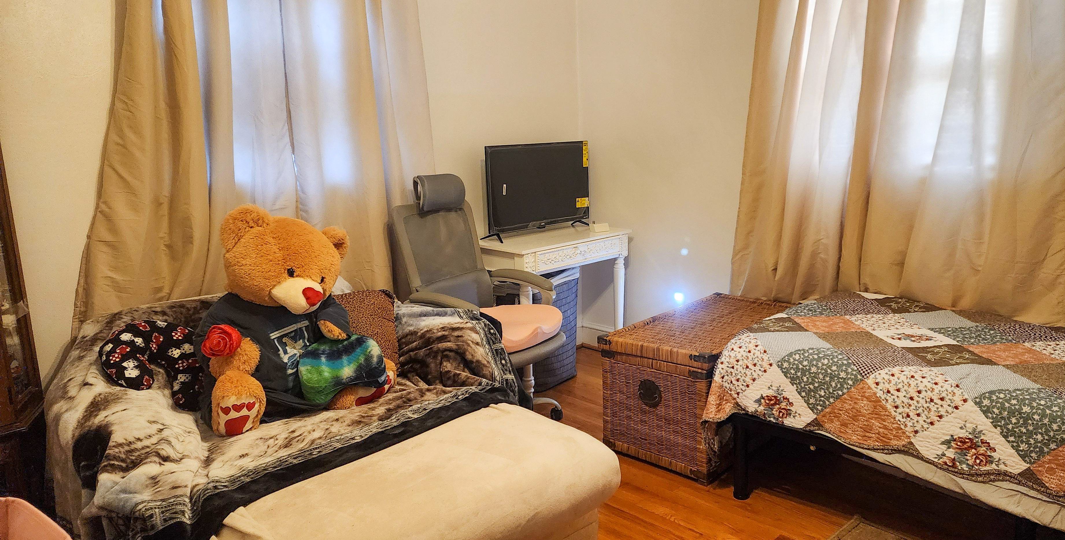 Cozy bedroom with a single bed, a small sofa holding a large teddy bear, a wicker chest, and a desk with a monitor near the window inside the home of Sponsored Residential Provider Cindy Freeman in Bassett, Virginia.