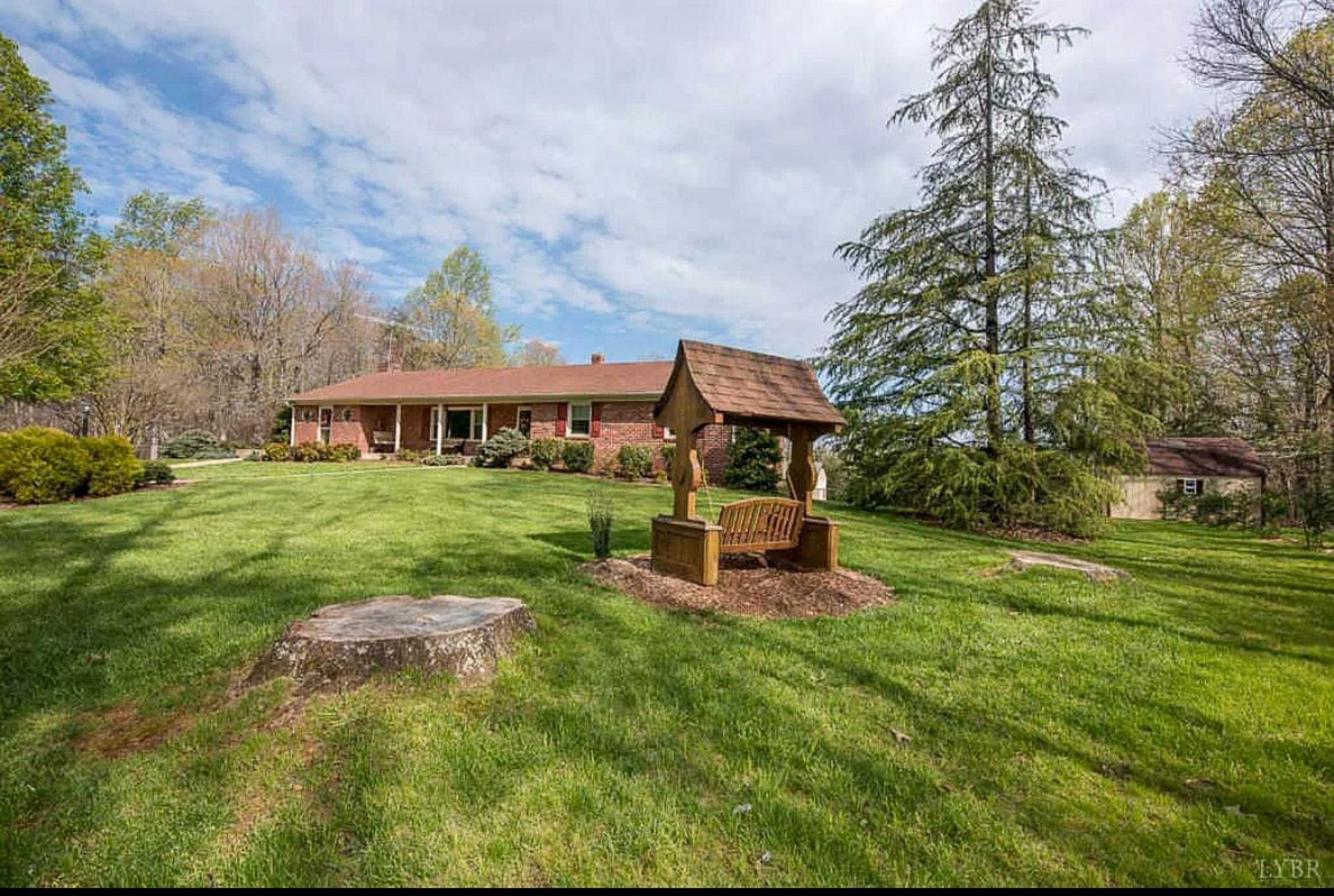 A large grassy backyard with a wooden wishing‑well structure in the center, surrounded by trees, with a brick ranch‑style house in the background at the home of Sponsored Residential Providers Carolyn and Phil Ware in Amherst, Virginia.