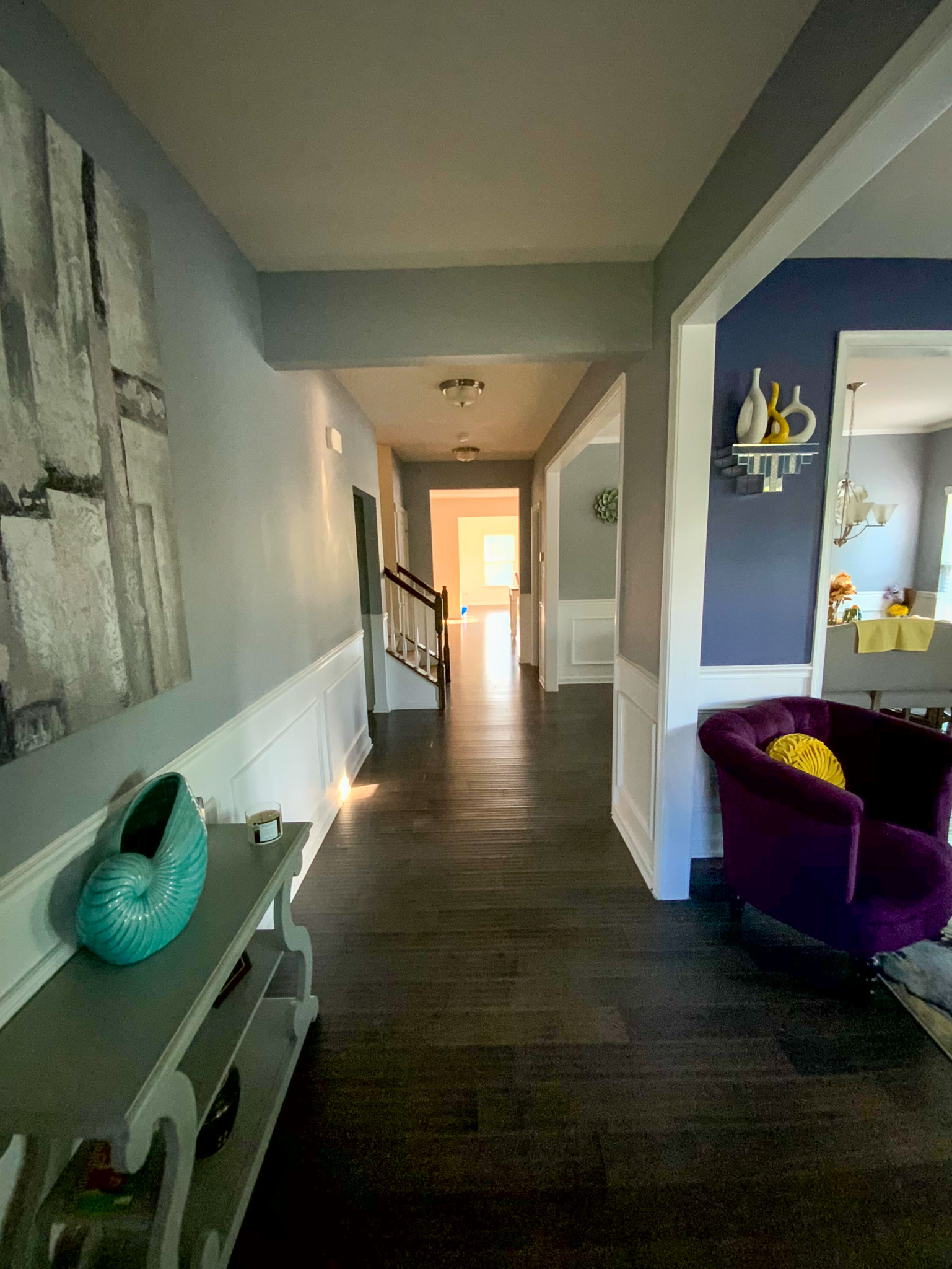 A long hallway with dark wood floors leads toward a bright room, with a glimpse of a purple chair and decorative accents in an adjacent space inside the home of Sponsored Residential Provider Derrick Moseley in Richmond, Virginia.