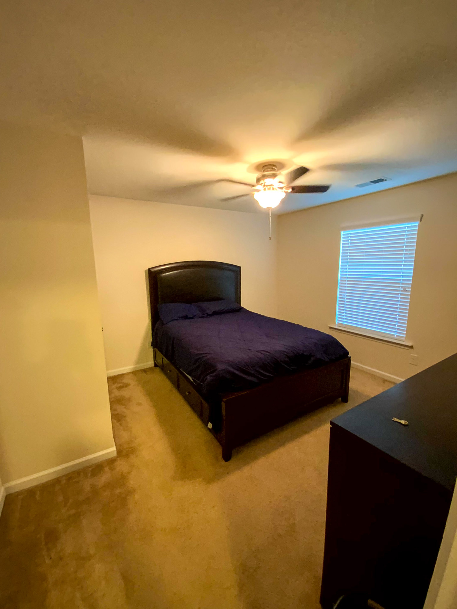 A simple bedroom contains a dark wood bed with navy bedding, a dresser, and a window that brings light into the space inside the home of Sponsored Residential Provider Derrick Moseley in Richmond, Virginia.