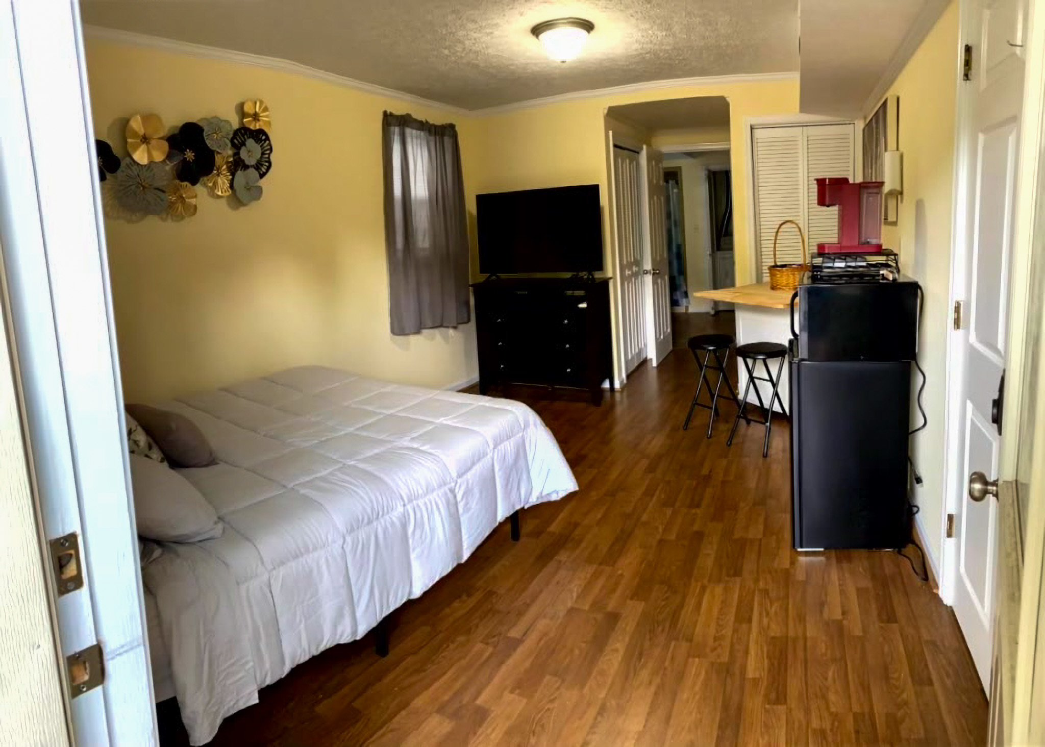 Room with a bed on the left, wood‑look flooring, a TV on a dark dresser, and a small kitchenette area with a counter and stools in the back inside the home of Sponsored Residential Providers Takeyshia Gibson and Antonio Carter in Forest, Virginia.