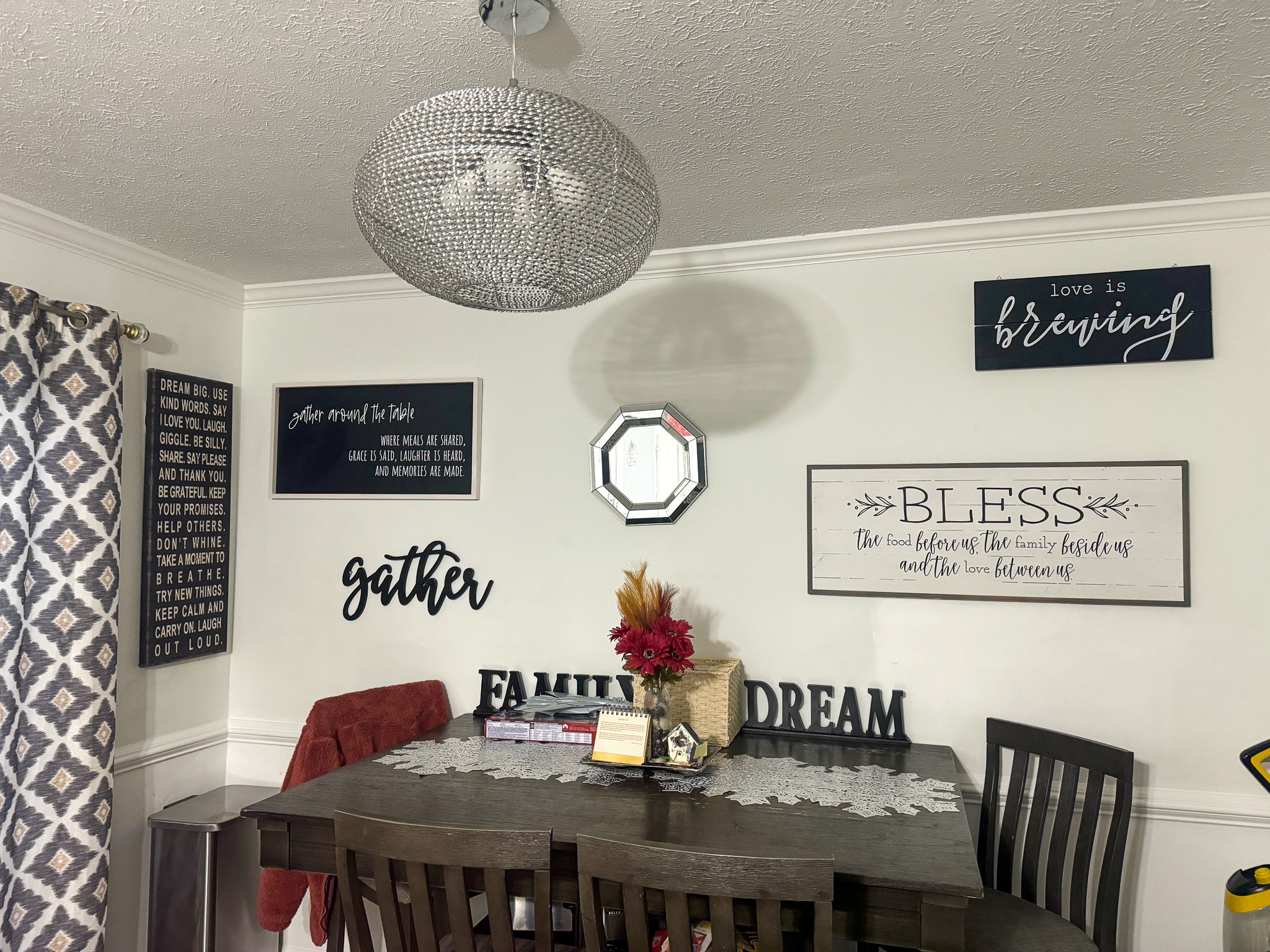 Dining area with a dark wooden table and chairs, decorative wall signs, patterned curtains, and a round mesh-style ceiling light inside the home of Sponsored Residential Providers Anthony Barnes and Demita Taylor in Forest, Virginia.