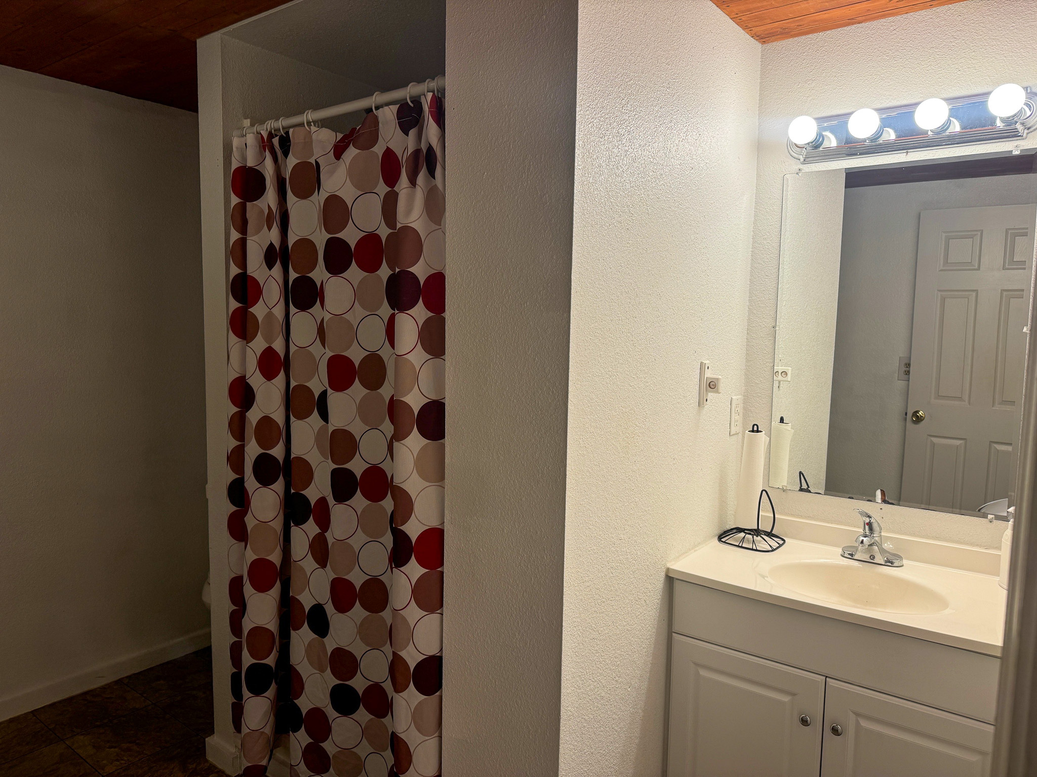 Bathroom with a white vanity and mirror on one side and a shower with a polka‑dot curtain on the other, under a wood-paneled ceiling inside the home of Sponsored Residential Providers Anthony Barnes and Demita Taylor in Forest, Virginia.