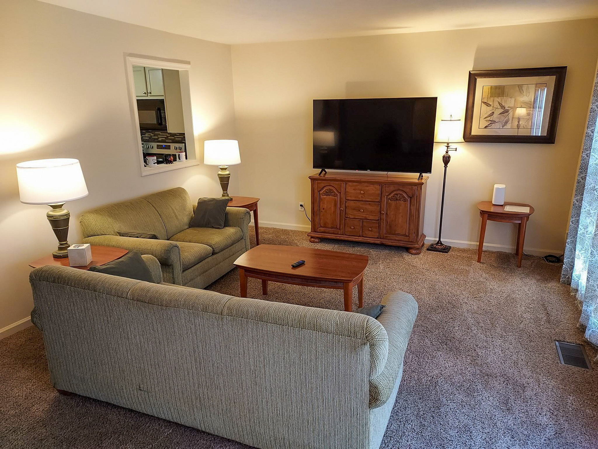 Living room with two green sofas, a wooden coffee table, lamps on end tables, and a large TV on a wooden stand inside the home of Sponsored Residential Provider David Wilcox in Harrisonburg, Virginia.