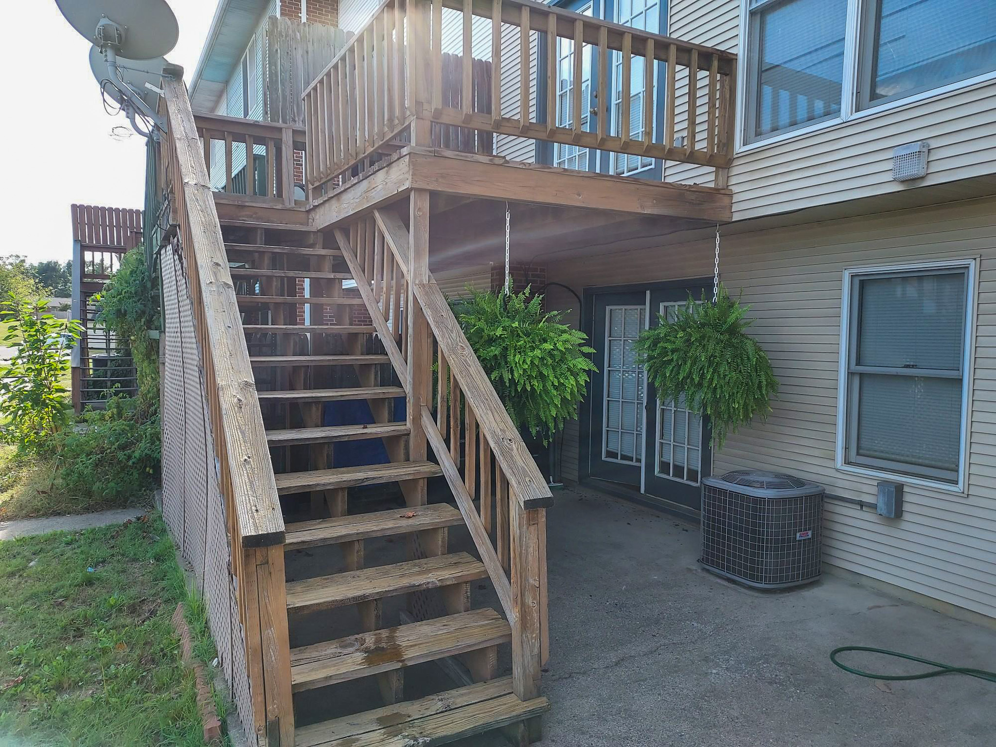 Exterior view of a home’s wooden deck and staircase leading to an upper level, with two hanging plants below and a sliding glass door on the lower patio at the home of Sponsored Residential Provider David Wilcox in Harrisonburg, Virginia.