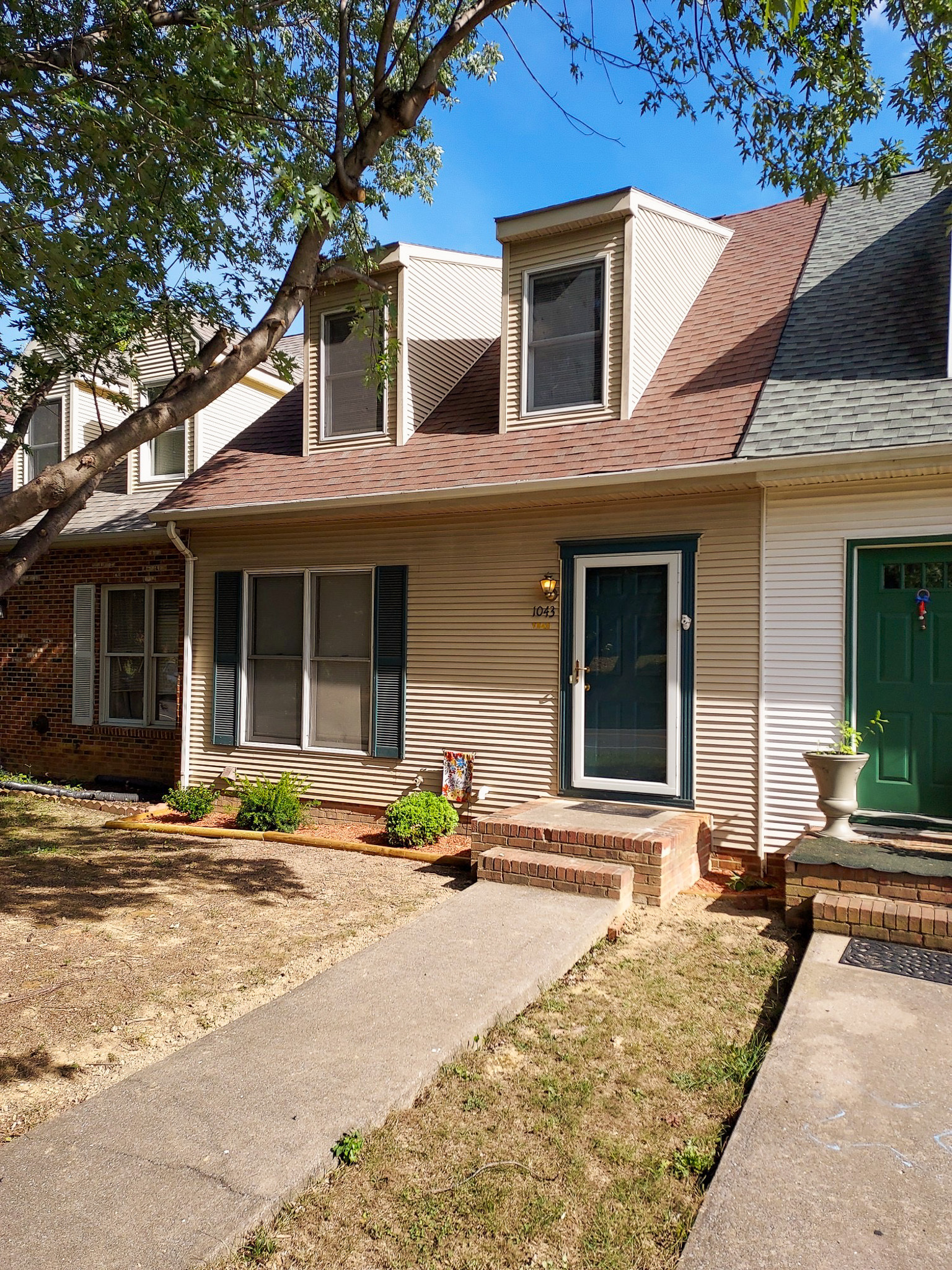 Townhouse with tan siding, a blue front door, dormer windows on the roof, and a small walkway leading to the entrance under a tree belonging to Sponsored Residential Provider David Wilcox in Harrisonburg, Virginia.
