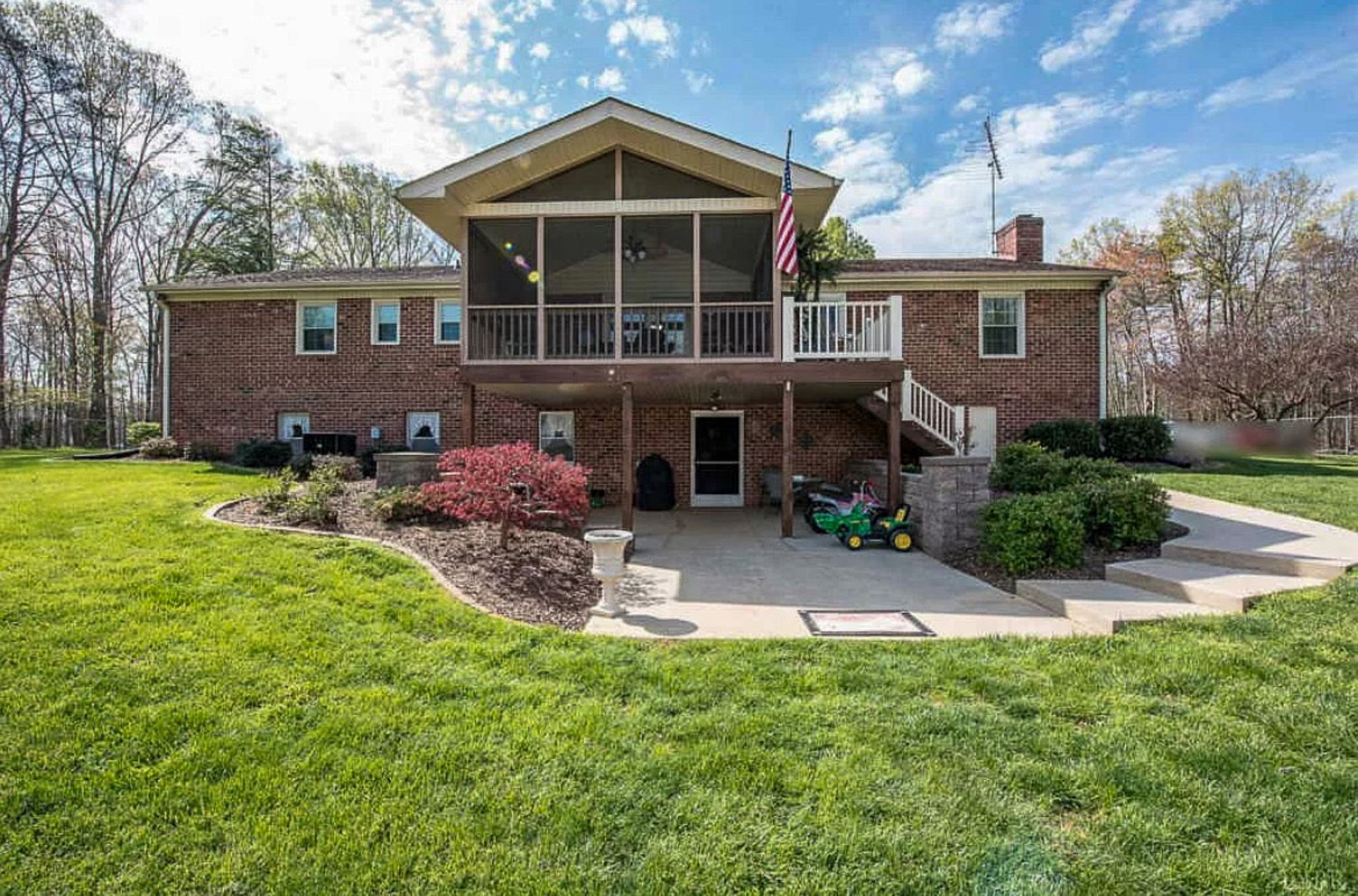 A brick house with a large covered upper deck and stairs leading down to a wide patio, surrounded by landscaped shrubs and an expansive green lawn at the home of Sponsored Residential Providers Carolyn and Phil Ware in Amherst, Virginia.