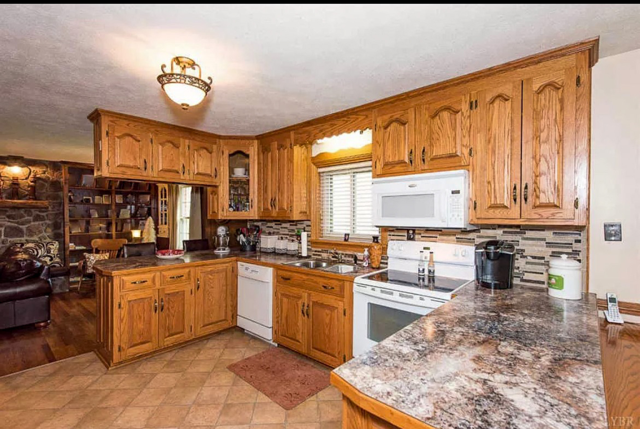 A kitchen with wooden cabinets, marble‑style countertops, white appliances, and a tile floor, opening into a living room with a stone fireplace inside the home of Sponsored Residential Providers Carolyn and Phil Ware in Amherst, Virginia.