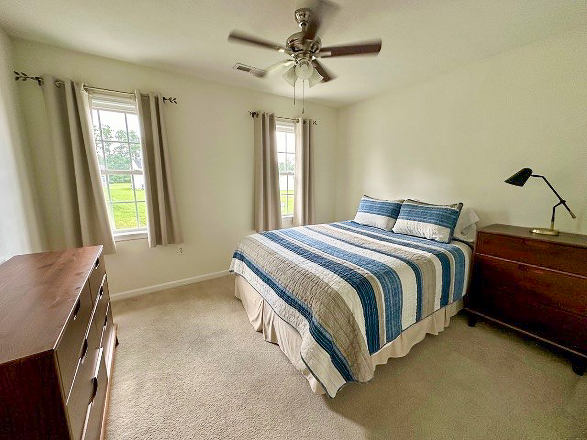Simple bedroom with a striped bedspread, two windows with beige curtains, wooden dressers, and a ceiling fan inside the home of Sponsored Residential Providers Nancy and Zach Kaplan in Waynesboro, Virginia.