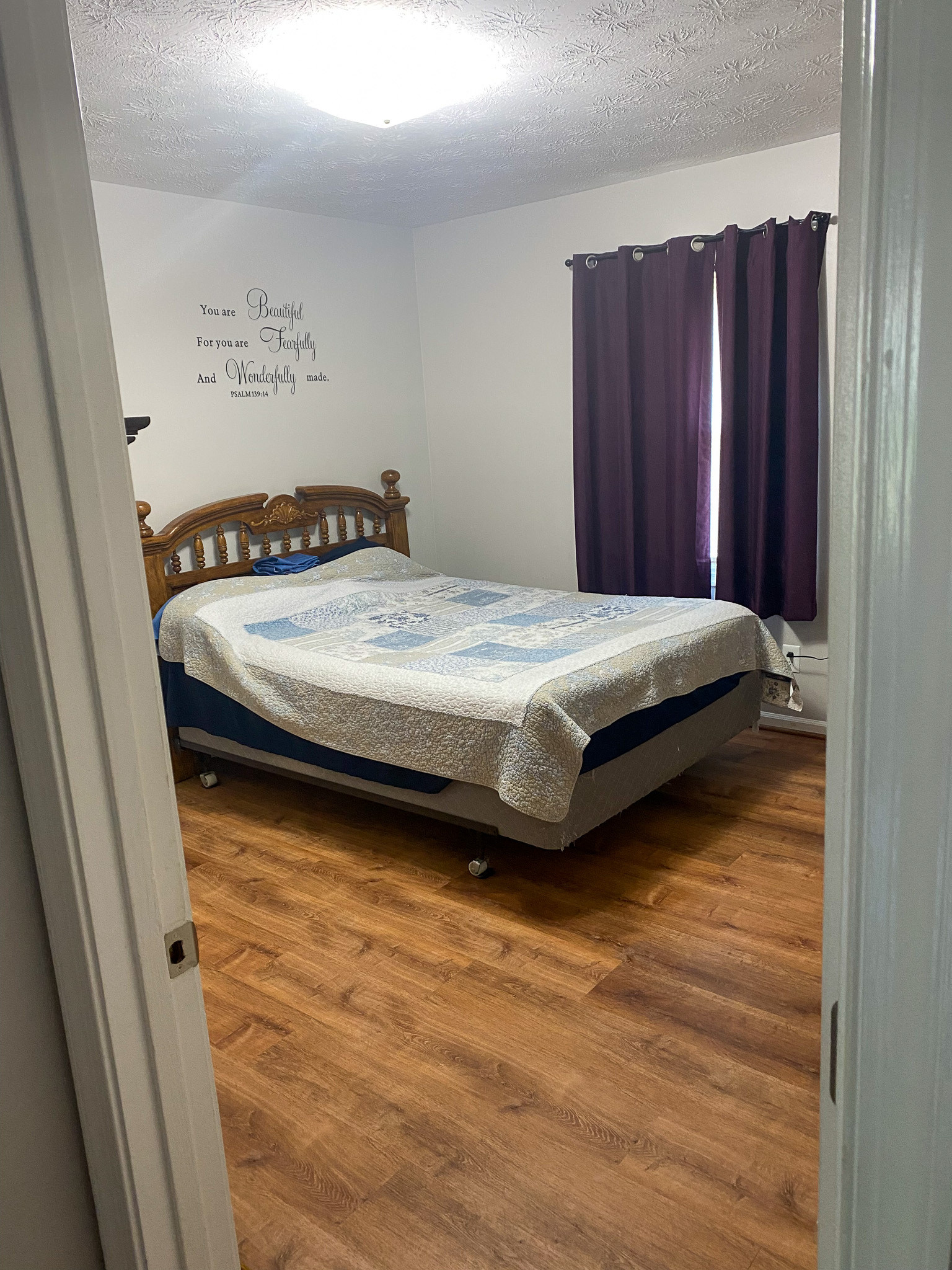 Bedroom with wood‑look flooring, a bed with a patterned quilt, a wooden headboard, white walls with decorative lettering, and dark curtains over the window inside the home of Sponsored Residential Providers Karen and Rob Robertson in Verona, Virginia.