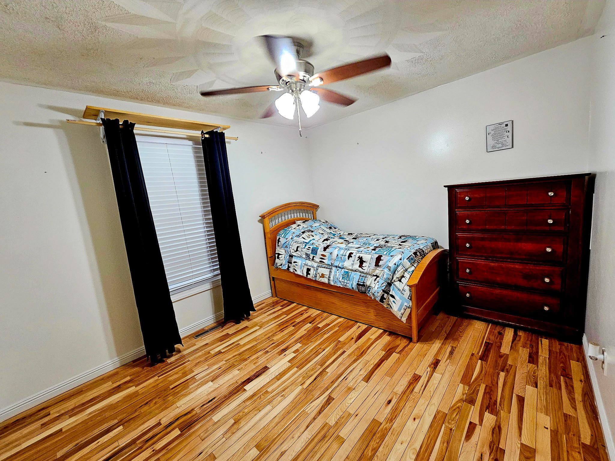 A simple bedroom with light wood floors, a single bed with a patterned quilt, dark curtains on a single window, a ceiling fan, and a tall wooden dresser against a plain wall inside the home of Group Home Provider Dylan Mays in Woodlawn, Virginia.