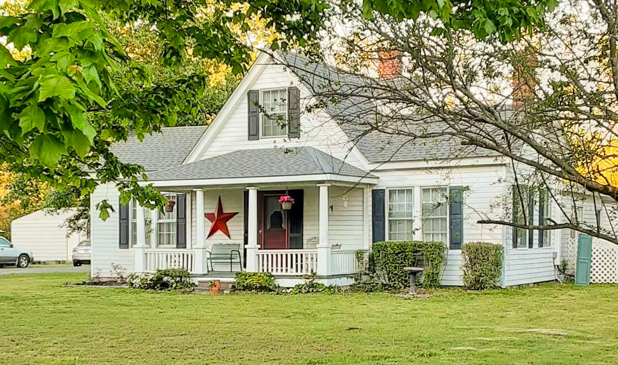 White farmhouse-style home with a front porch, dark shutters, and a large red star on the siding, set on a grassy lawn with trees framing the view belonging to Sponsored Residential Provider Angela Justice in Mathews, Virginia.
