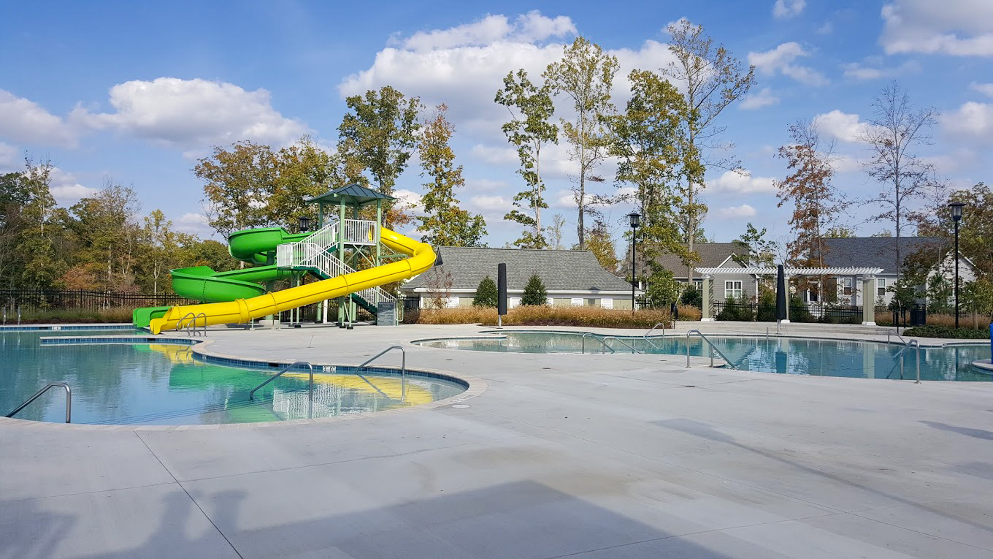 An outdoor pool area features a large green and yellow waterslide beside wide concrete decking and surrounding buildings in the community of Sponsored Residential Provider Derrick Moseley in Richmond, Virginia.