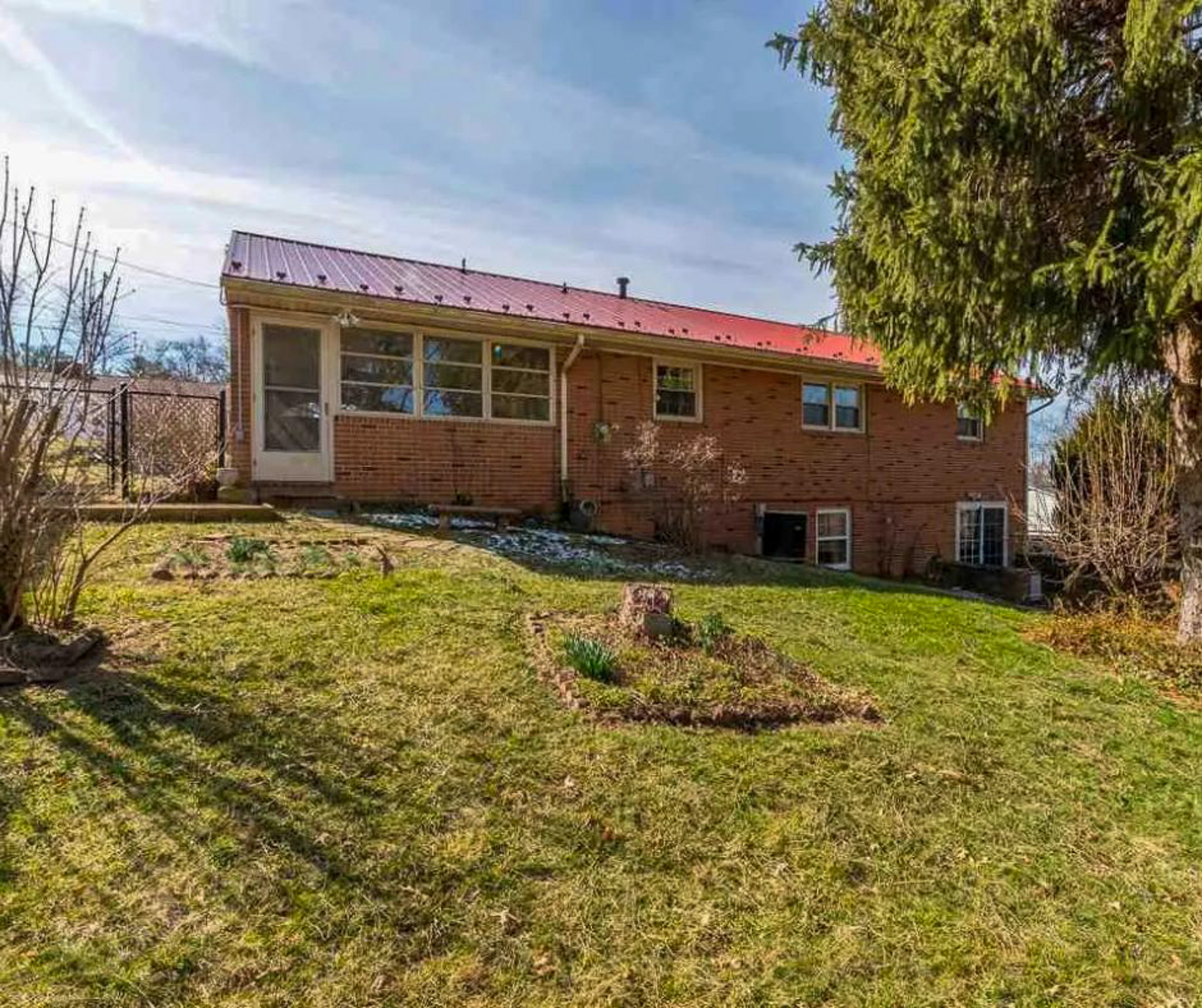 Rear view of a brick house with a red metal roof, enclosed porch area, basement windows, and a spacious grassy yard with a large tree and small garden bed belonging to Sponsored Residential Providers Bill and Heather Terry in Staunton, Virginia.