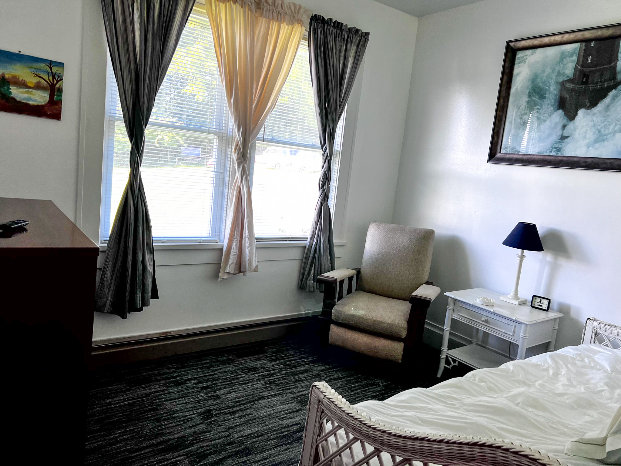 Bedroom with large windows and sheer curtains, an armchair, bedside table with lamp, and a bed partially visible in the foreground inside the home of Sponsored Residential Provider Tammy Hunt in Floyd, Virginia.