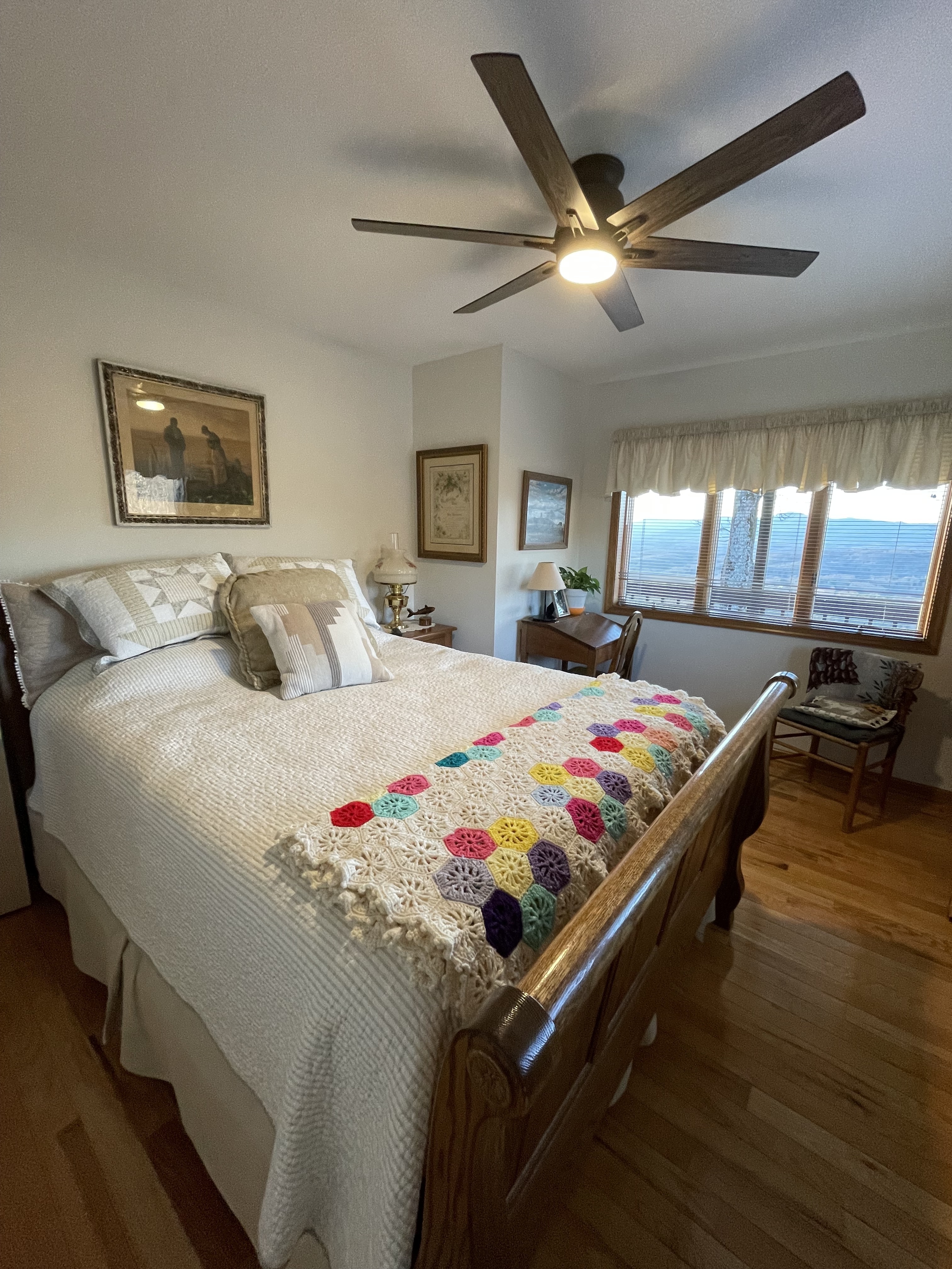 Bedroom with a large wooden bed covered in white bedding and a colorful crocheted throw, ceiling fan above, wall art, and a window with mountain views inside the home of Sponsored Residential Providers Jimmy and Nancy Ayers in Monroe, Virginia.