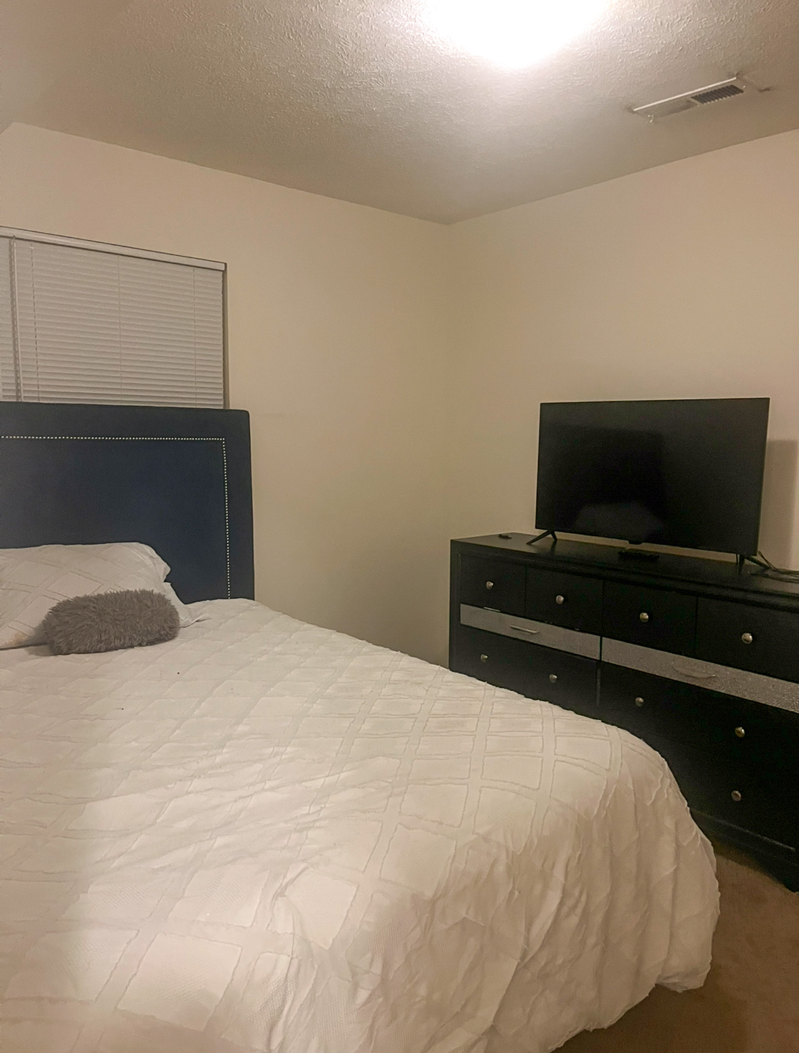 Bedroom with a white bedspread, dark headboard, and a dresser holding a TV against a beige wall inside the home of Sponsored Residential Provider Kanesha Moseley in Lynchburg, Virginia.