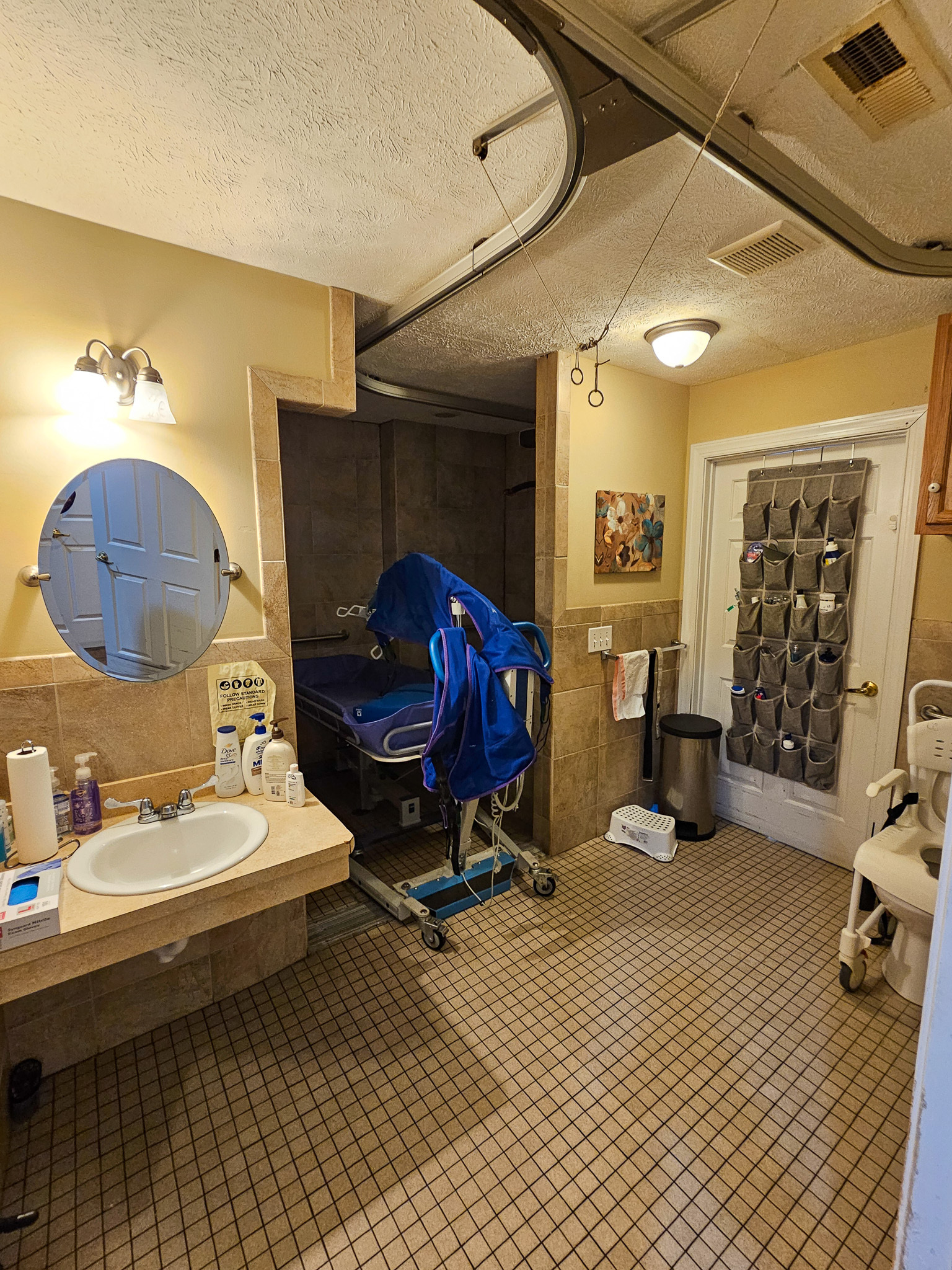 A bathroom with yellow walls features a sink and mirror on the left, a medical lift device near the shower area, and a patterned glass door on the right at Boonsboro Group Home in Lynchburg, Virginia.