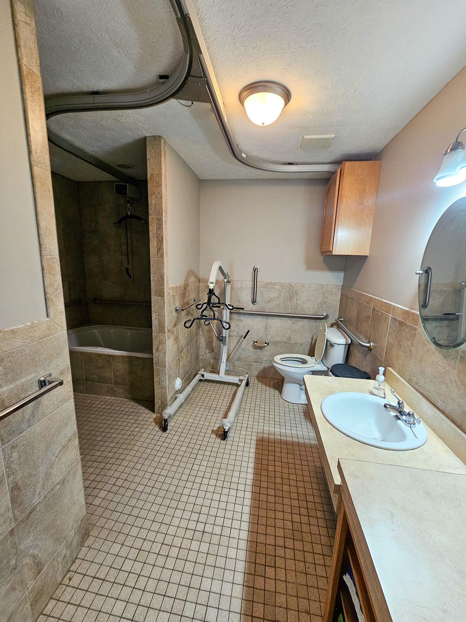 A bathroom with tiled floors and walls contains a vanity, toilet, walk‑in shower, and a medical lift device near the center of the room at Boonsboro Group Home in Lynchburg, Virginia.
