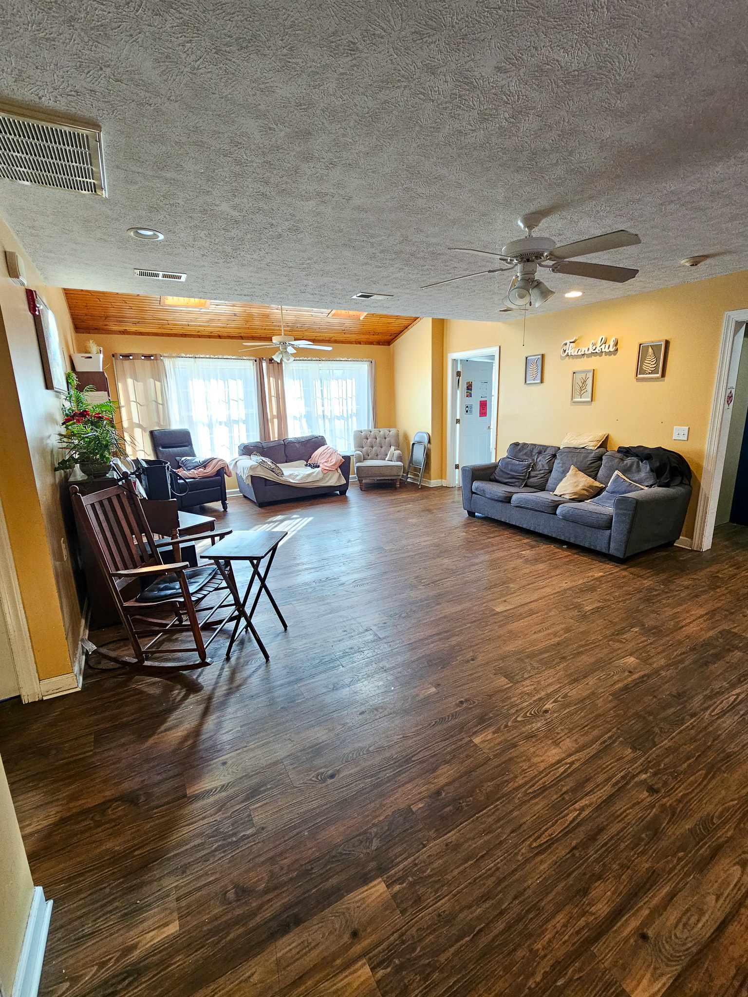 A spacious living area with dark wood floors features a gray sofa, rocking chairs, and additional seating near large windows at Boonsboro Group Home in Lynchburg, Virginia.