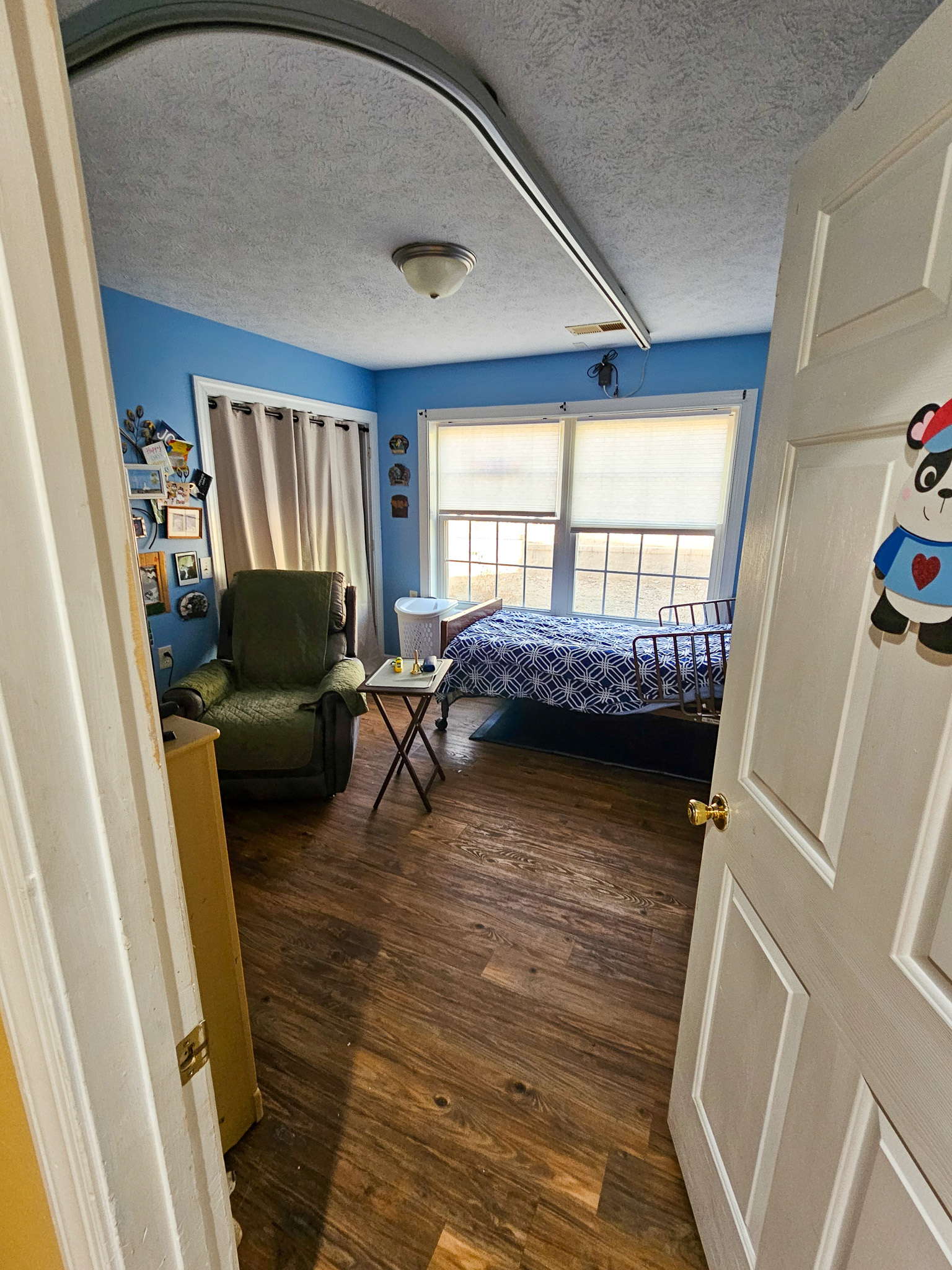 A bedroom with blue walls contains a bed by the window, a green recliner, and a ceiling‑mounted lift track at Boonsboro Group Home in Lynchburg, Virginia.