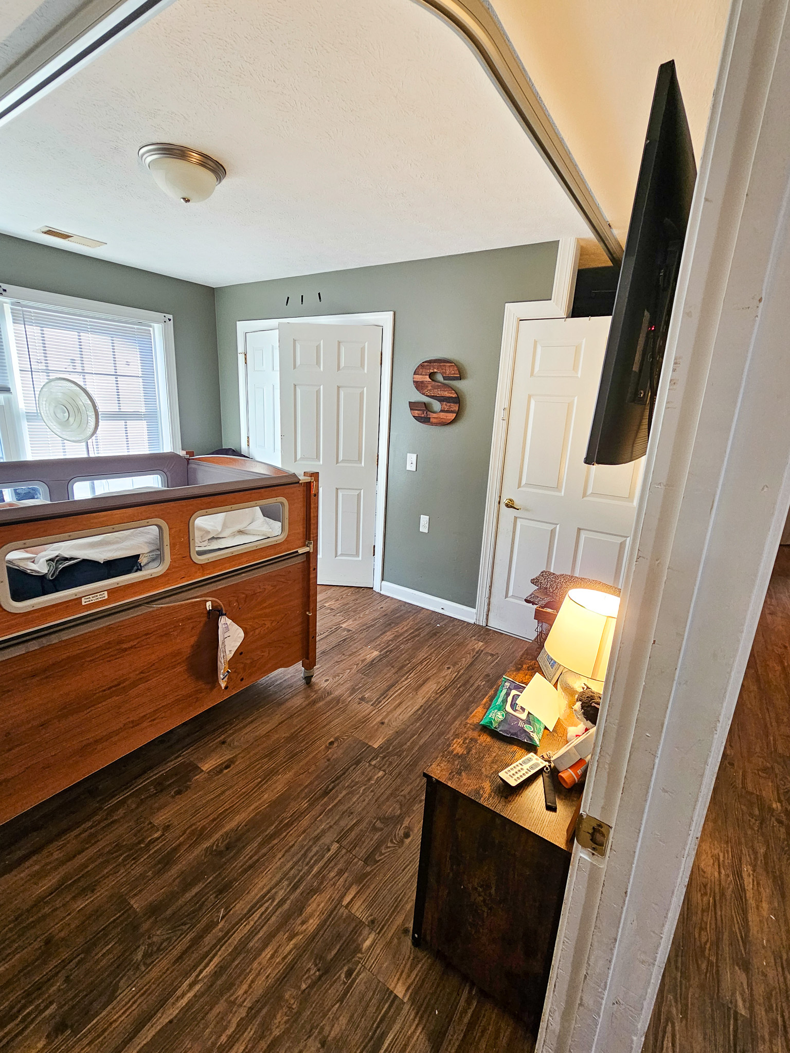 A bedroom with green walls features a hospital‑style bed, wood furniture, and a ceiling‑mounted lift track at Boonsboro Group Home in Lynchburg, Virginia.