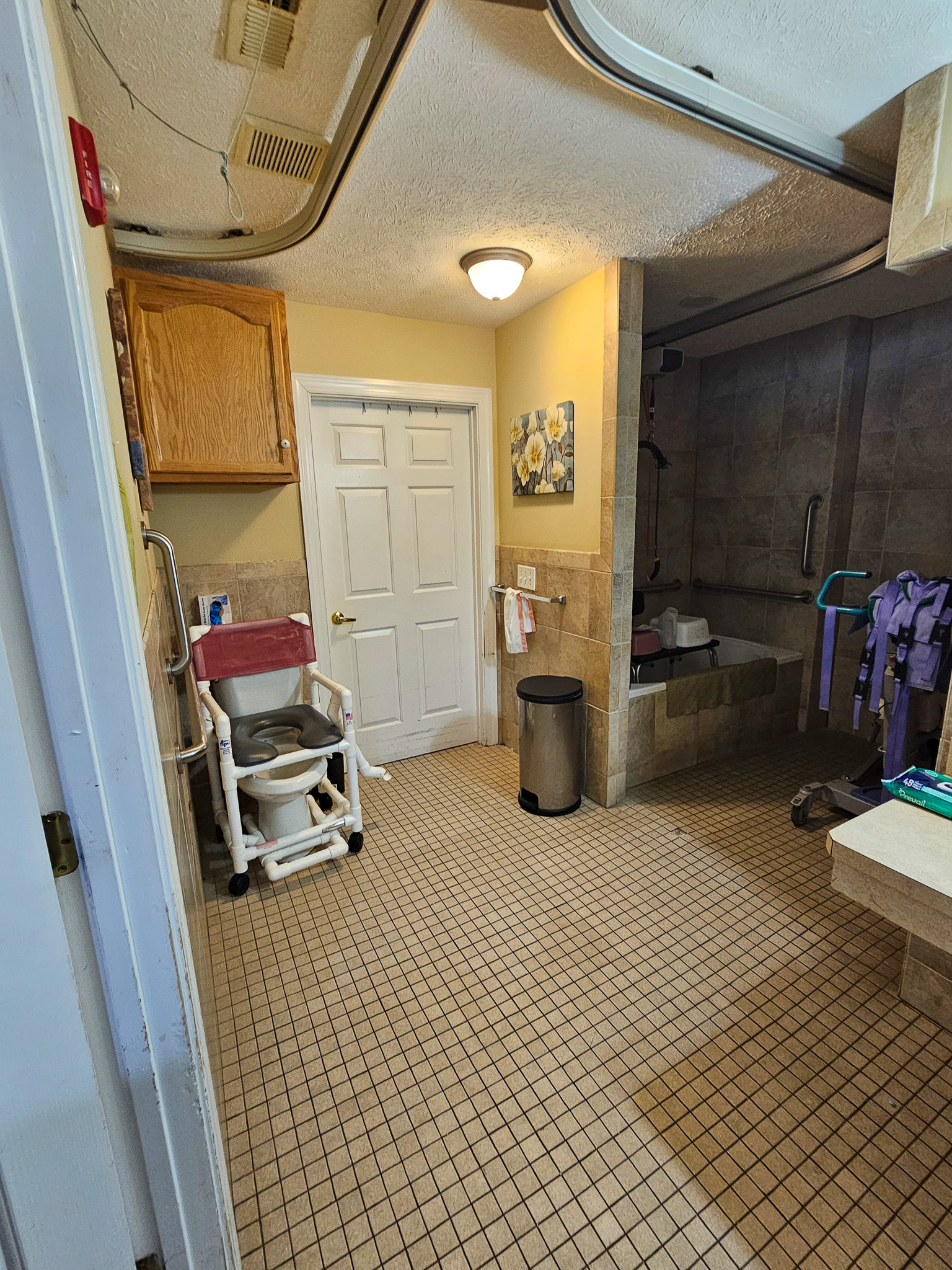 A tiled bathroom with yellow walls contains a shower area, a sink counter, storage cabinets, and a shower chair near the doorway at Boonsboro Group Home in Lynchburg, Virginia.