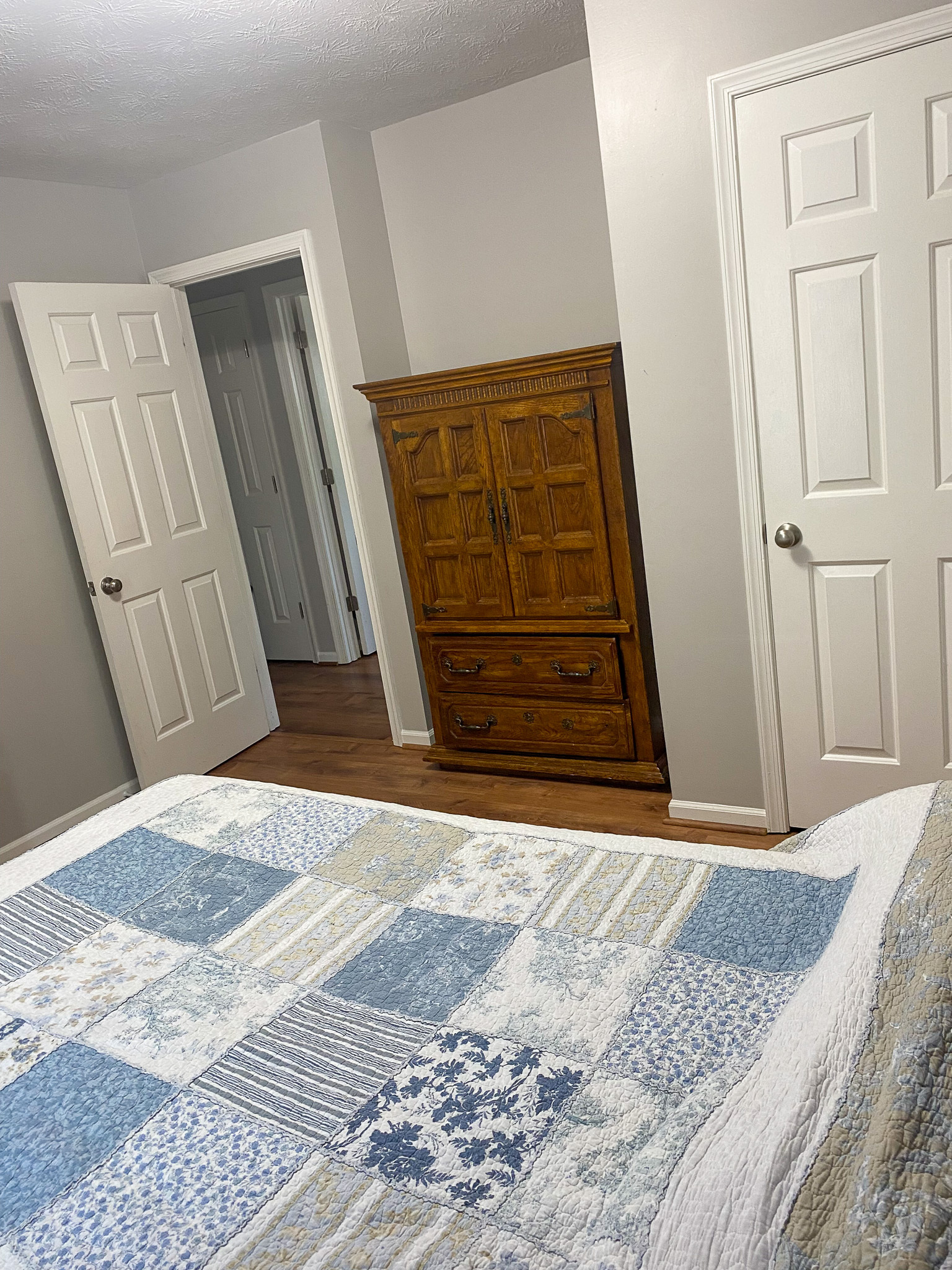 Bedroom view showing a bed with a blue and white patchwork quilt, wood‑look flooring, a wooden armoire, and two white doors—one open to a hallway and one closed inside the home of Sponsored Residential Providers Karen and Rob Robertson in Verona, Virginia.