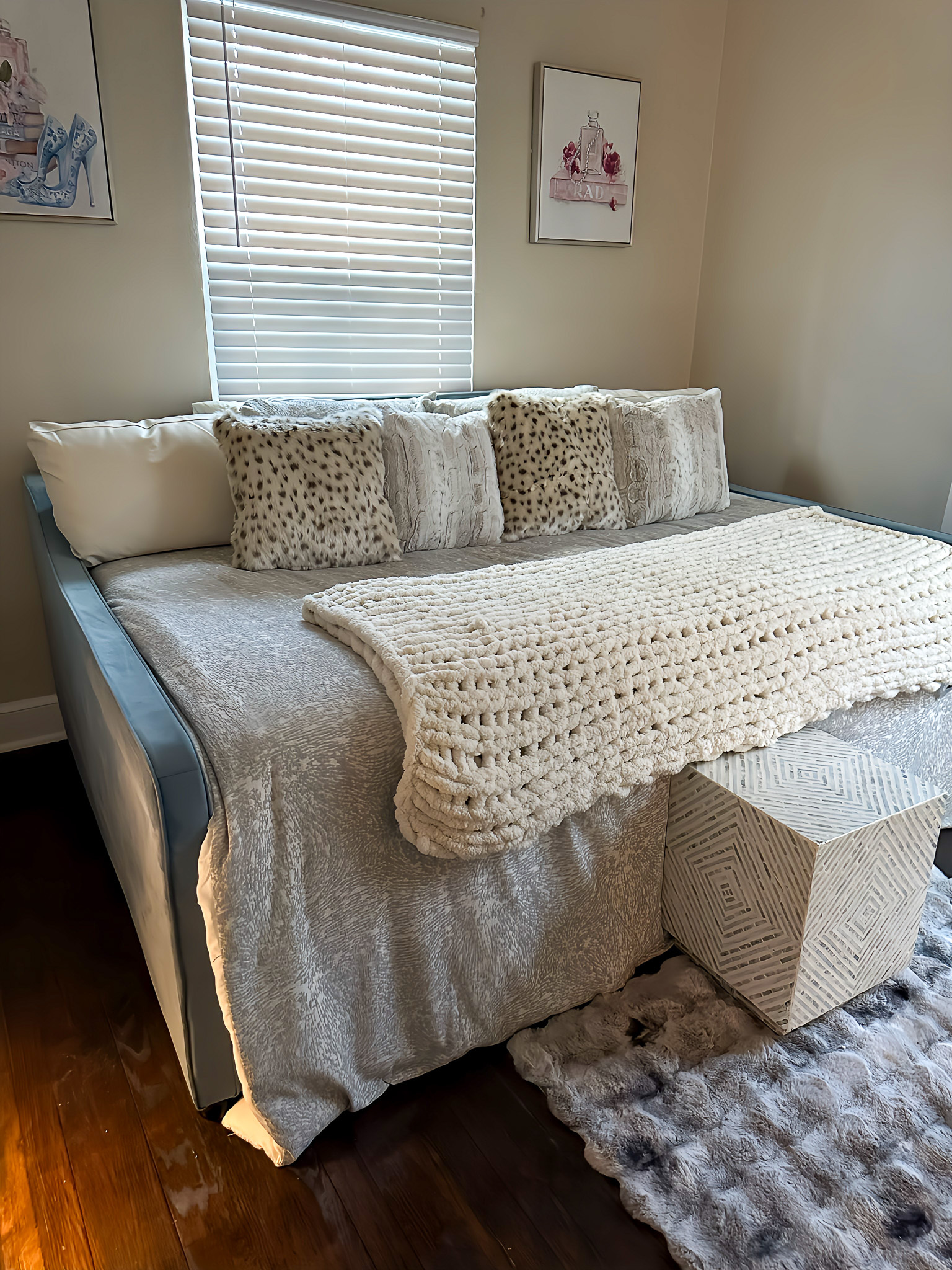 A cozy bedroom with a neatly made bed topped with textured pillows and a knitted blanket beside a small cube ottoman inside the home of Sponsored Residential Providers Gia & Celia Calloway in Roanoke, Virginia.