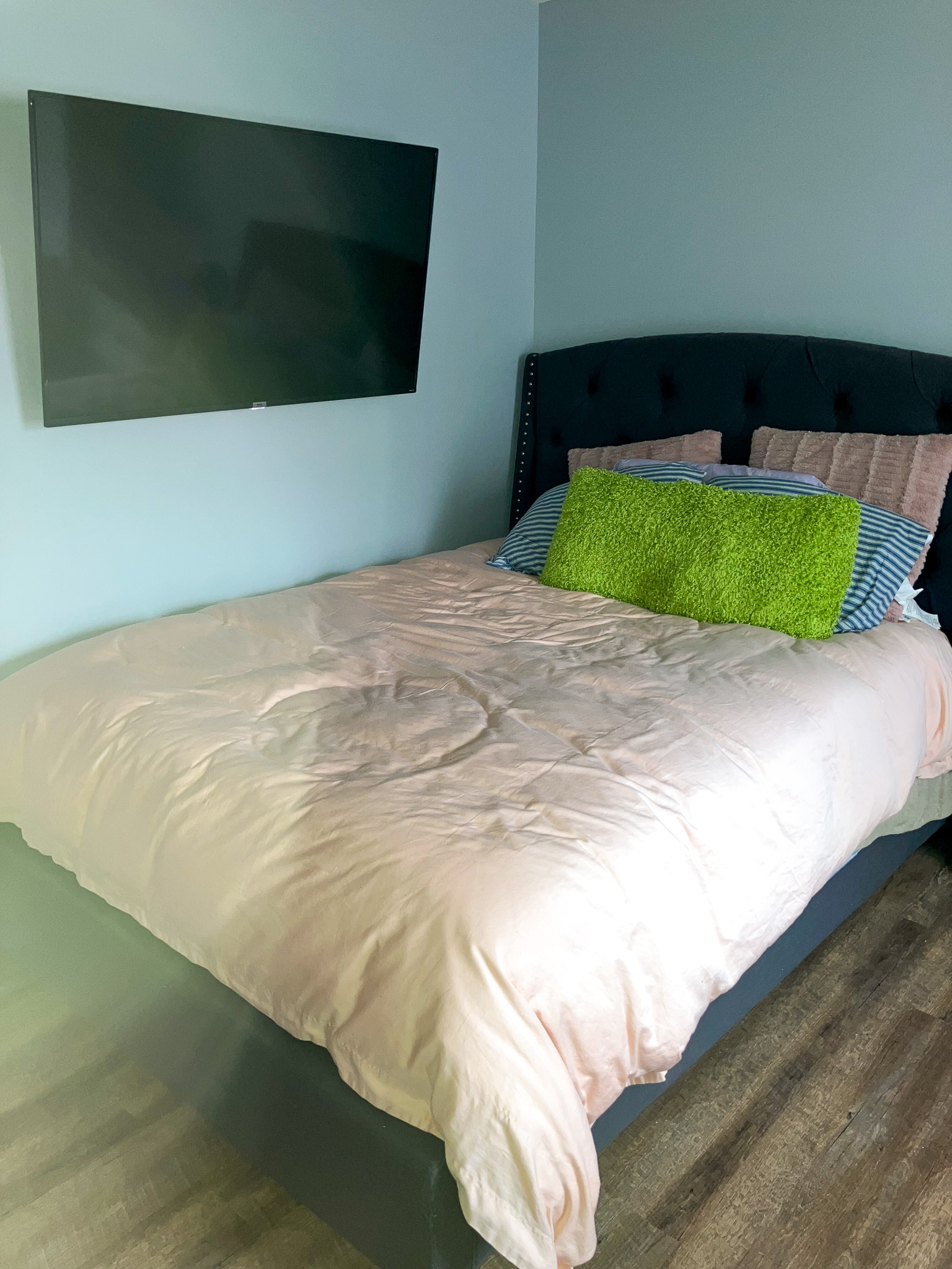 Bedroom with a wall‑mounted TV and a bed with light bedding and colorful pillows on wood‑style flooring inside the home of Sponsored Residential Providers Crystal and Christopher Cameron in Fredericksburg, Virginia.