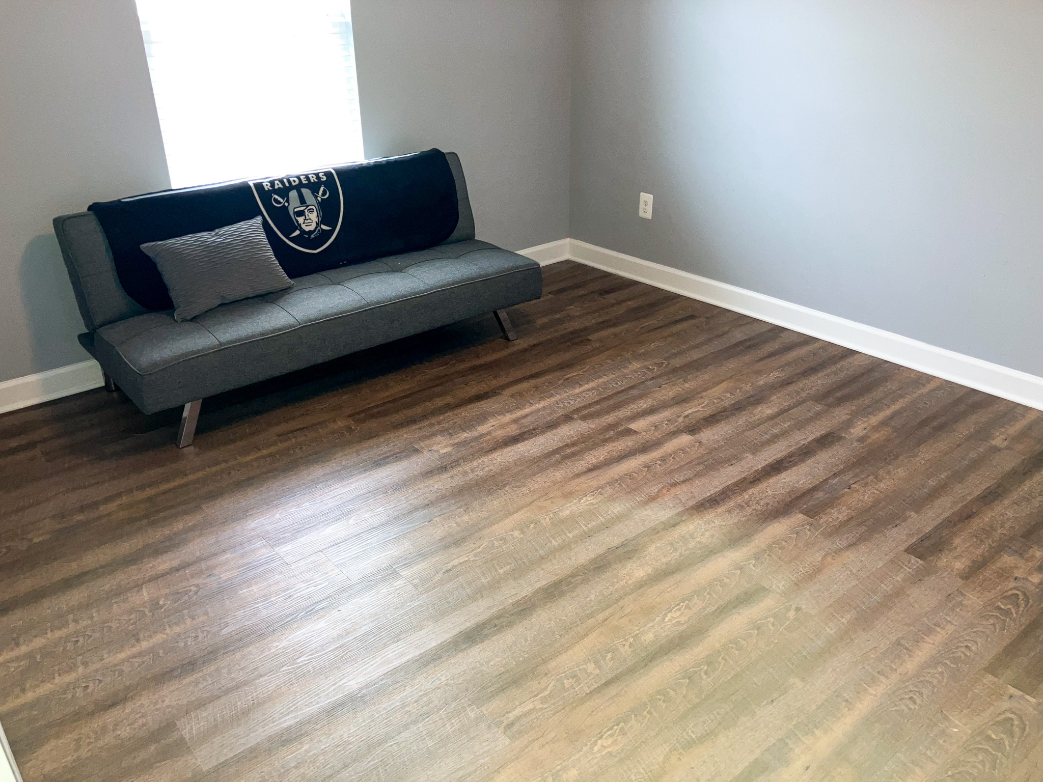 Simple room with gray walls, wood‑style flooring, and a gray futon near a window inside the home of Sponsored Residential Providers Crystal and Christopher Cameron in Fredericksburg, Virginia.