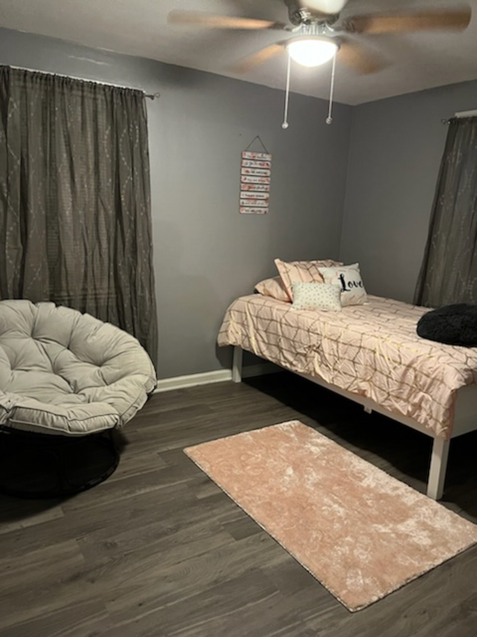 A bedroom with a single bed, a round cushioned chair, dark curtains, and a small pink rug on the floor inside the home of Sponsored Residential Provider Claudia Davis in Roanoke, Virginia.