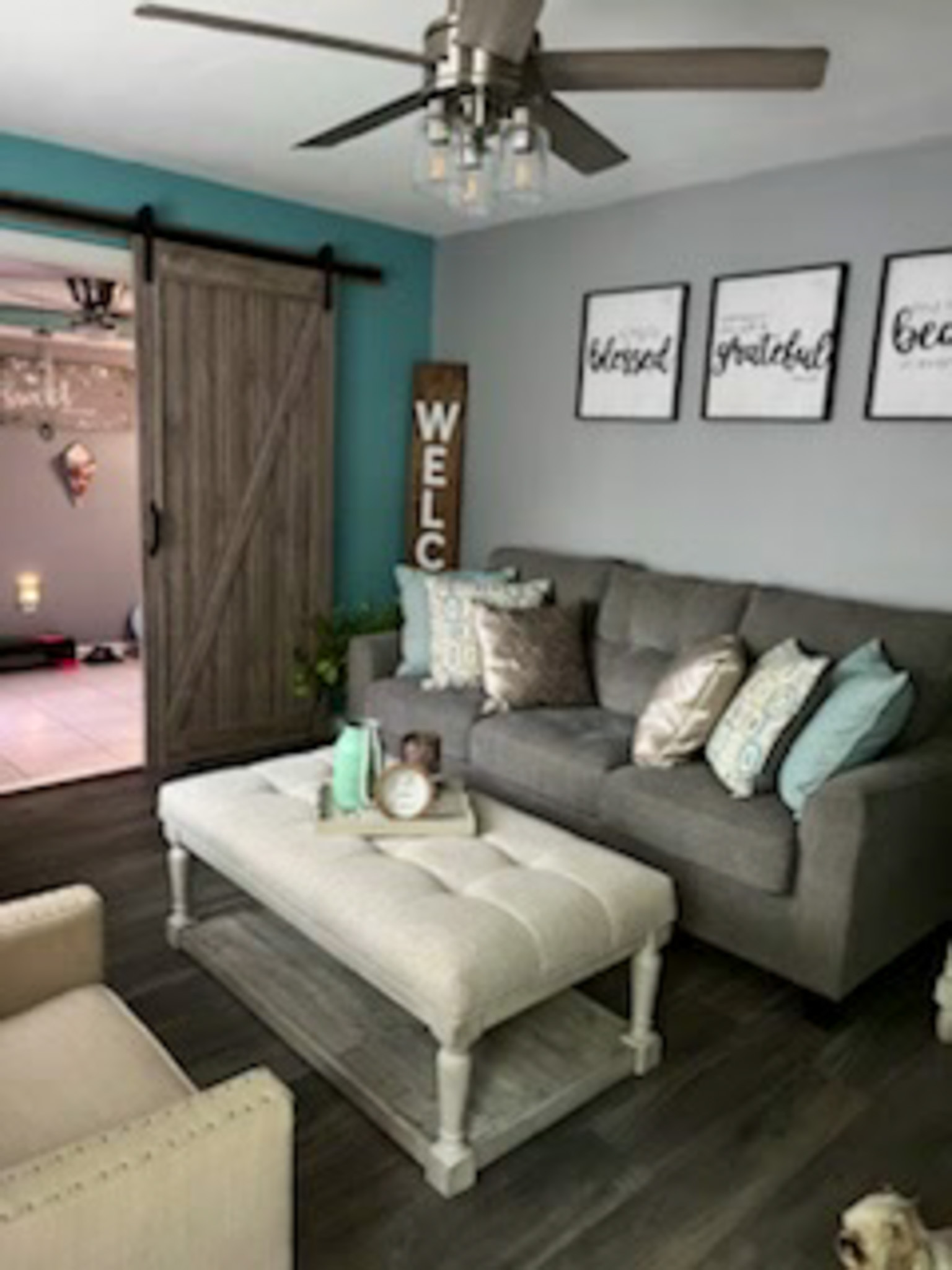 A living room with a gray sofa, an upholstered coffee table, and a sliding barn door against a teal accent wall inside the home of Sponsored Residential Provider Claudia Davis in Roanoke, Virginia.