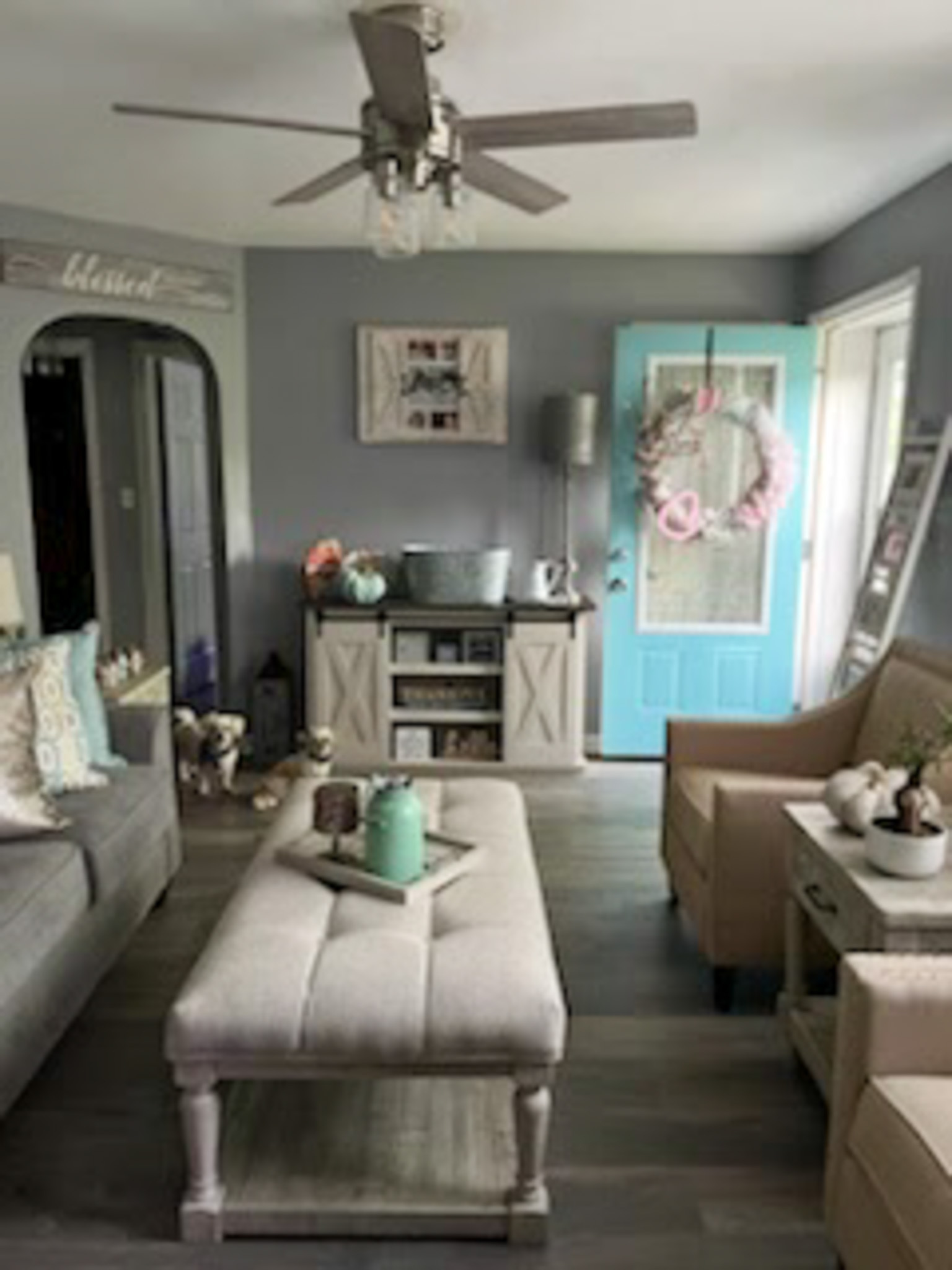 A cozy living room with gray walls, neutral furniture, a long upholstered coffee table, and a bright turquoise door decorated with a floral wreath inside the home of Sponsored Residential Provider Claudia Davis in Roanoke, Virginia.