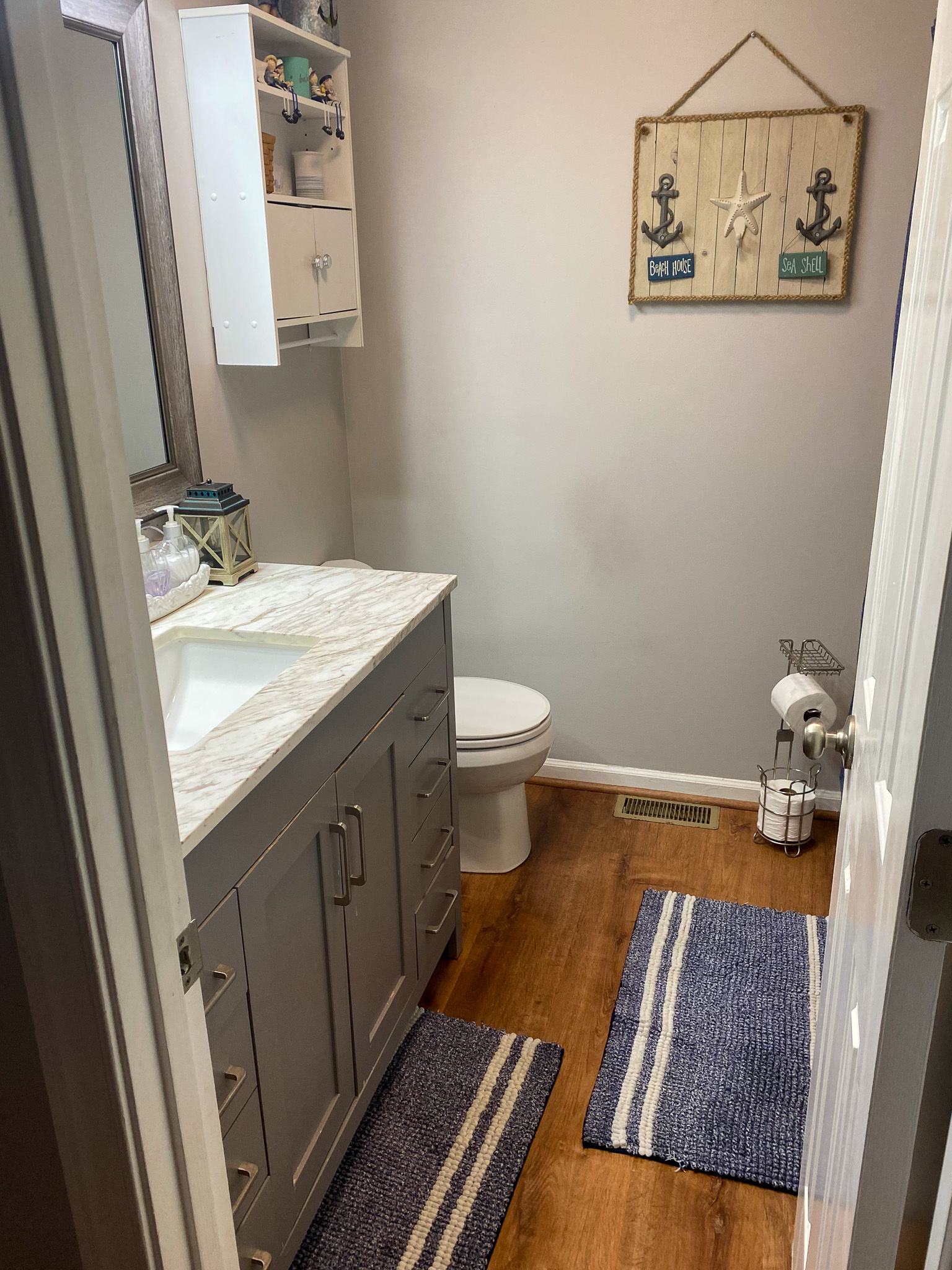 Bathroom with a gray vanity and white countertop, wood‑look flooring, toilet, wall‑mounted cabinet, and nautical-themed decor on a light-colored wall inside the home of Sponsored Residential Providers Karen and Rob Robertson in Verona, Virginia.