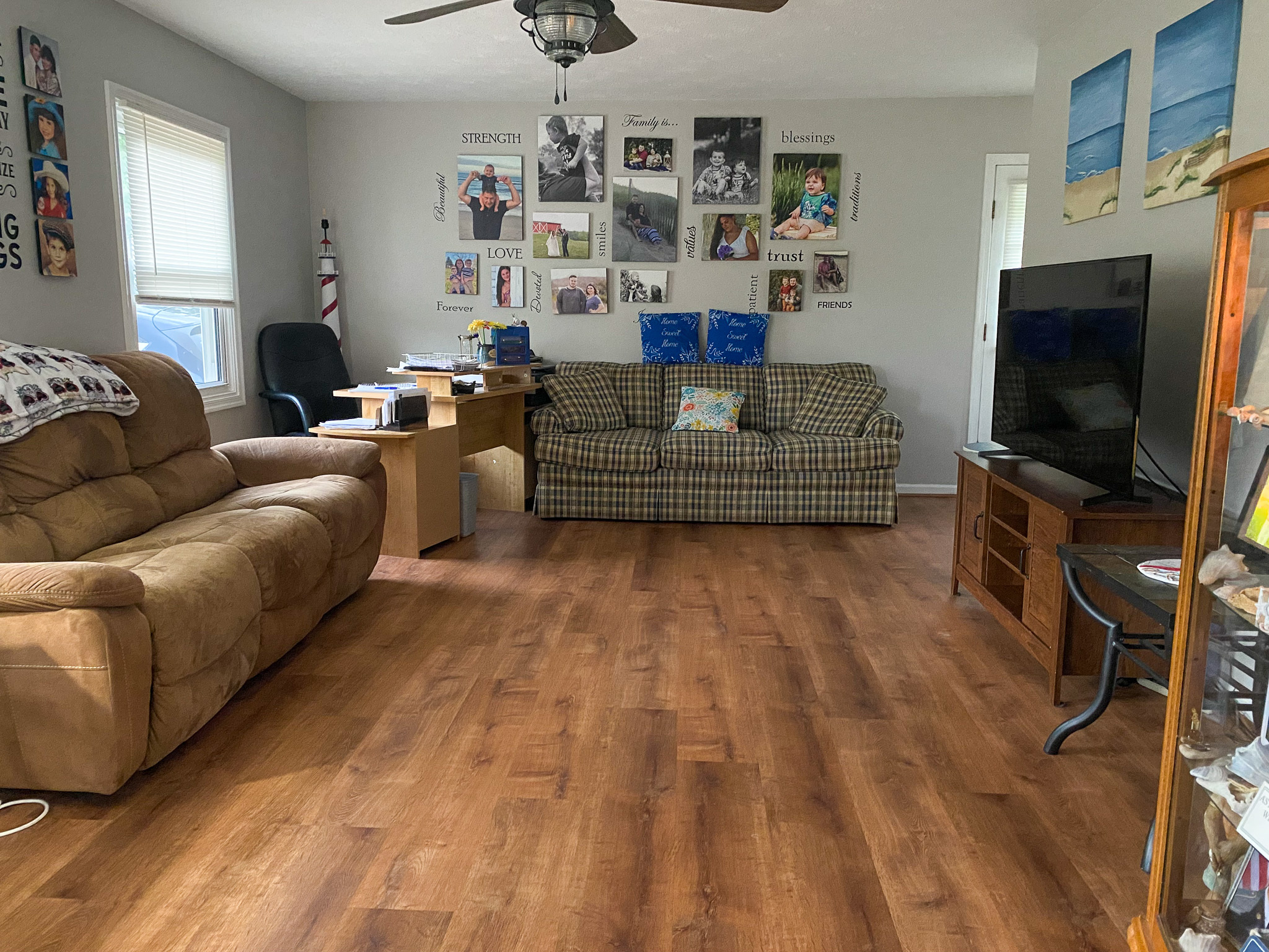 Living room with wood‑look flooring, a brown recliner, plaid sofa, wall of framed photos, a desk and office chair, ceiling fan, and a TV on a stand inside the home of Sponsored Residential Providers Karen and Rob Robertson in Verona, Virginia.
