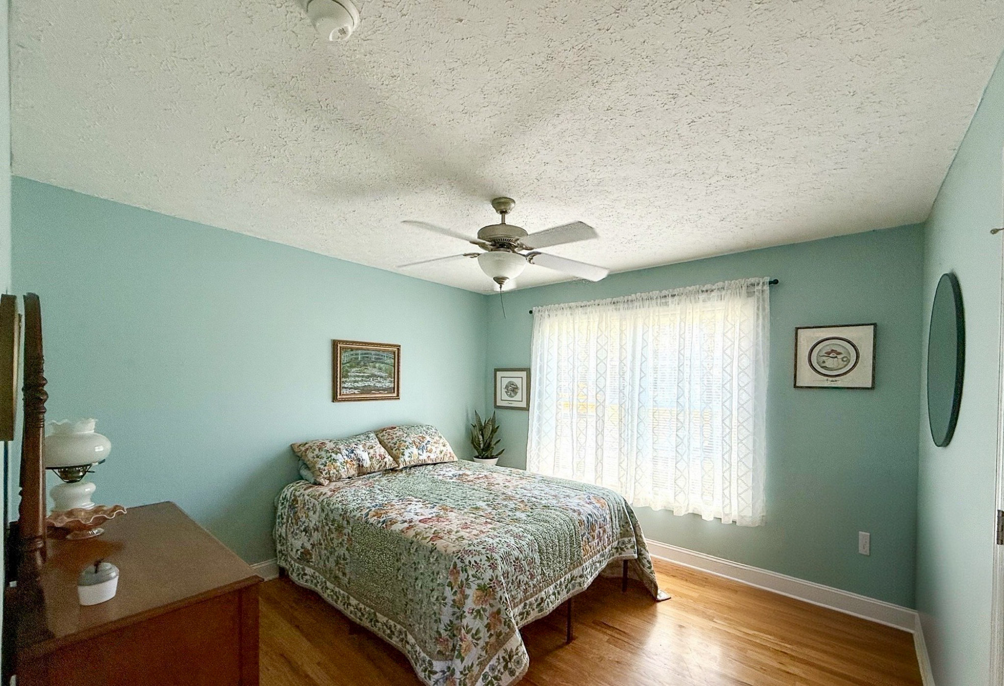 A bedroom with light green walls, a patterned bedspread, a wooden dresser, sheer curtains, and a ceiling fan in the home of Group Home Providers Kim and Rick Duncan in Patrick Springs, Virginia.