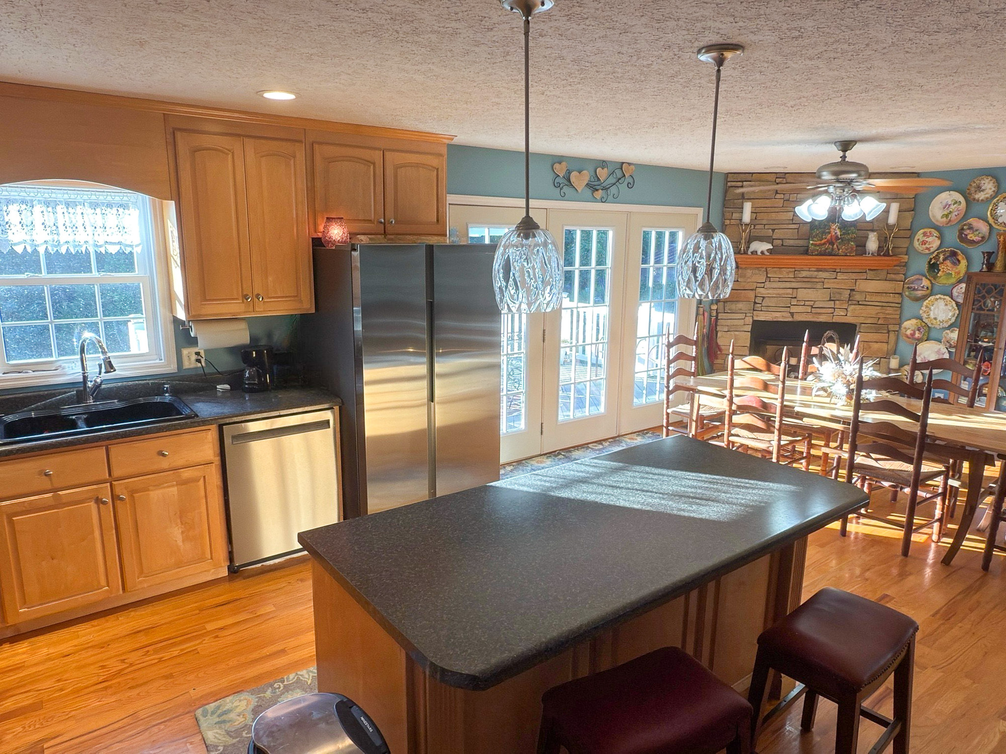 A kitchen with wooden cabinets, a central island, stainless steel appliances, and double doors leading to a dining area with a stone fireplace inside the home of Group Home Providers Kim and Rick Duncan in Patrick Springs, Virginia.