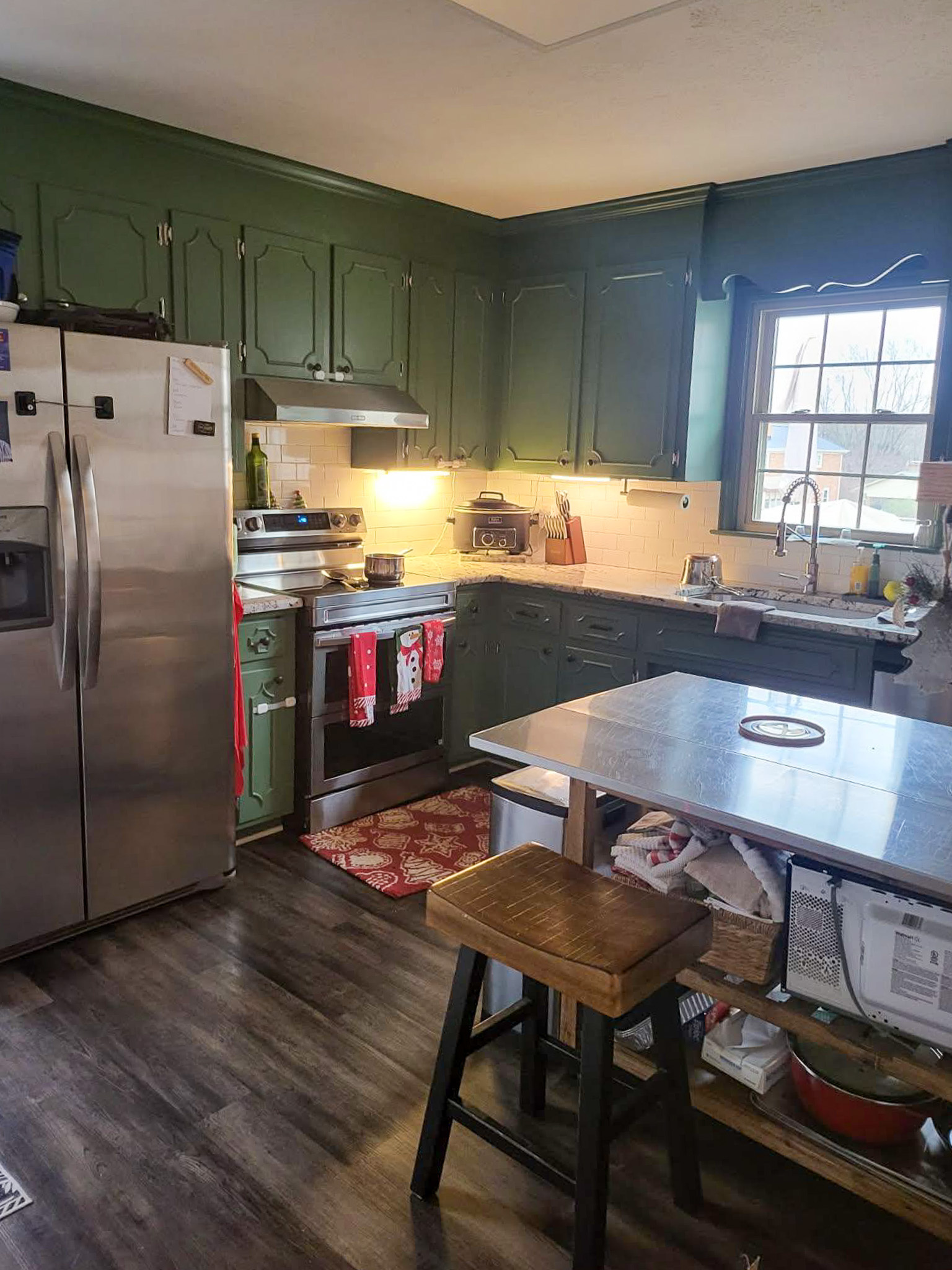 A kitchen with green cabinets features stainless steel appliances, a window over the sink, and a small island with seating inside the home of Sponsored Residential Providers Scott and Tedra Flanders in Lynchburg, Virginia.