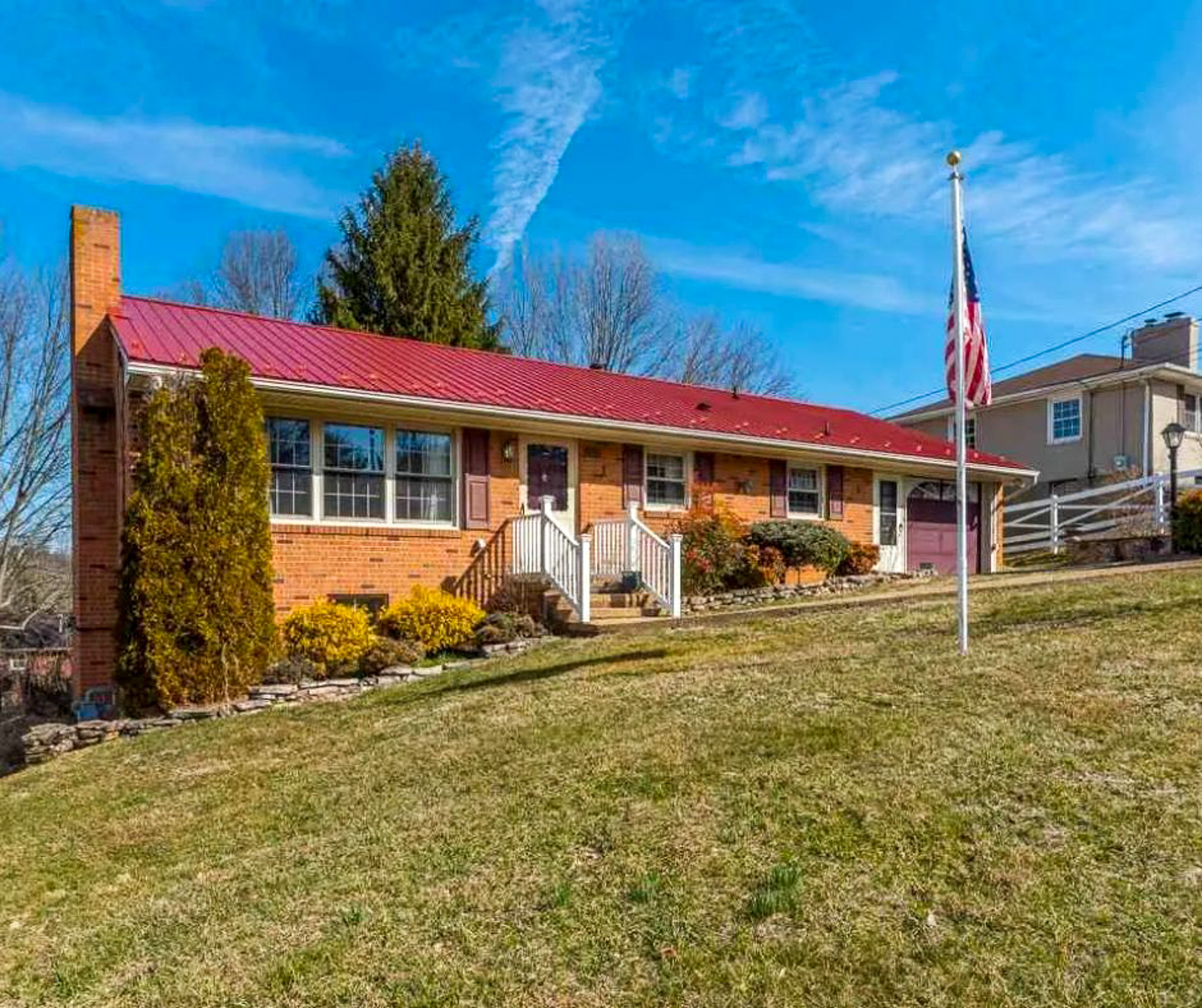 Brick ranch‑style house with a red metal roof, front steps leading to the entry, landscaped shrubs, and a flagpole on a grassy hill belonging to Sponsored Residential Providers Bill and Heather Terry in Staunton, Virginia.
