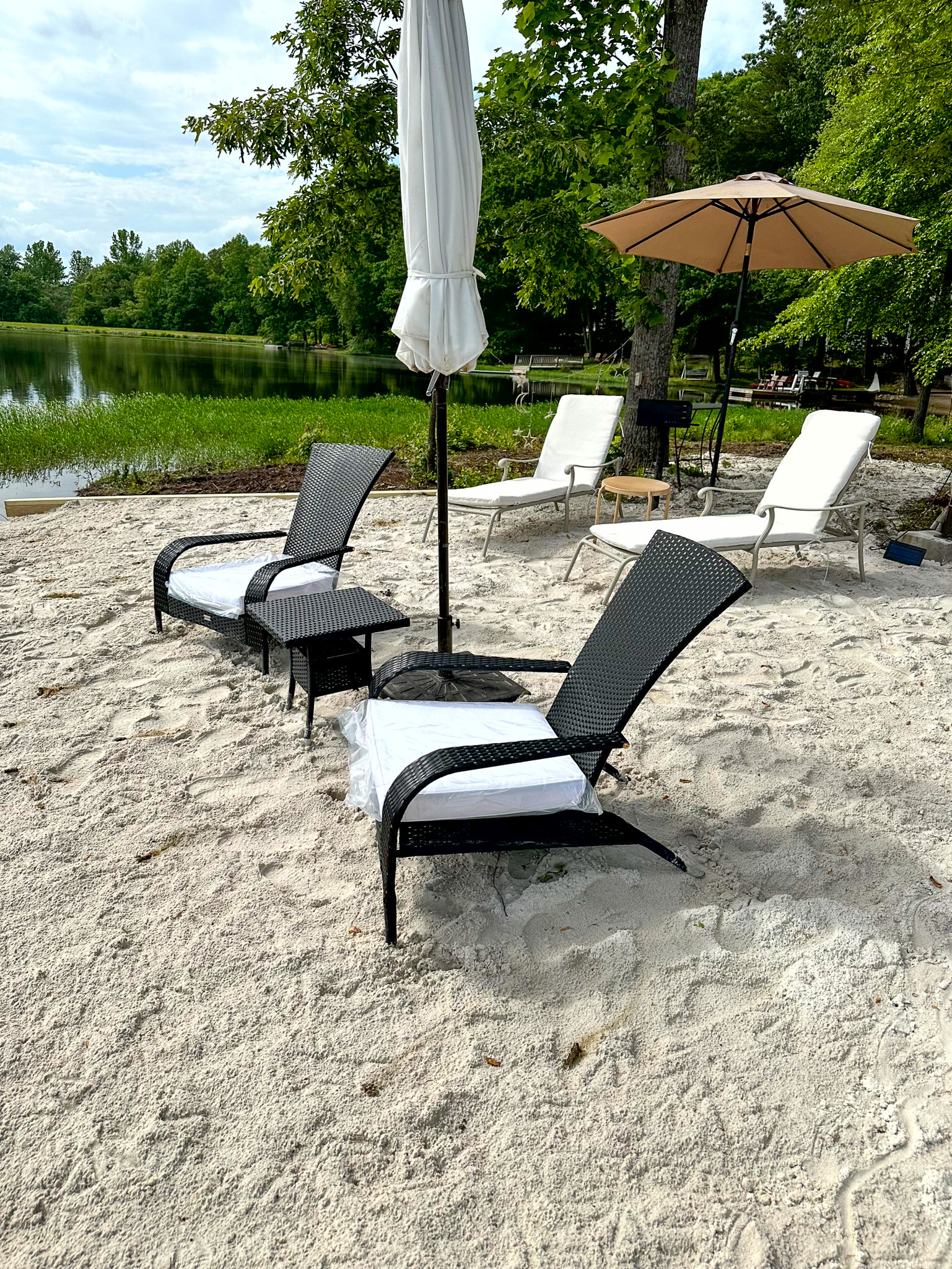 Lounge chairs and umbrellas set on a sandy lakeside beach with green trees and calm water in the background outside the home of Group Home Provider Ana Garcia in Spotsylvania, Virginia.