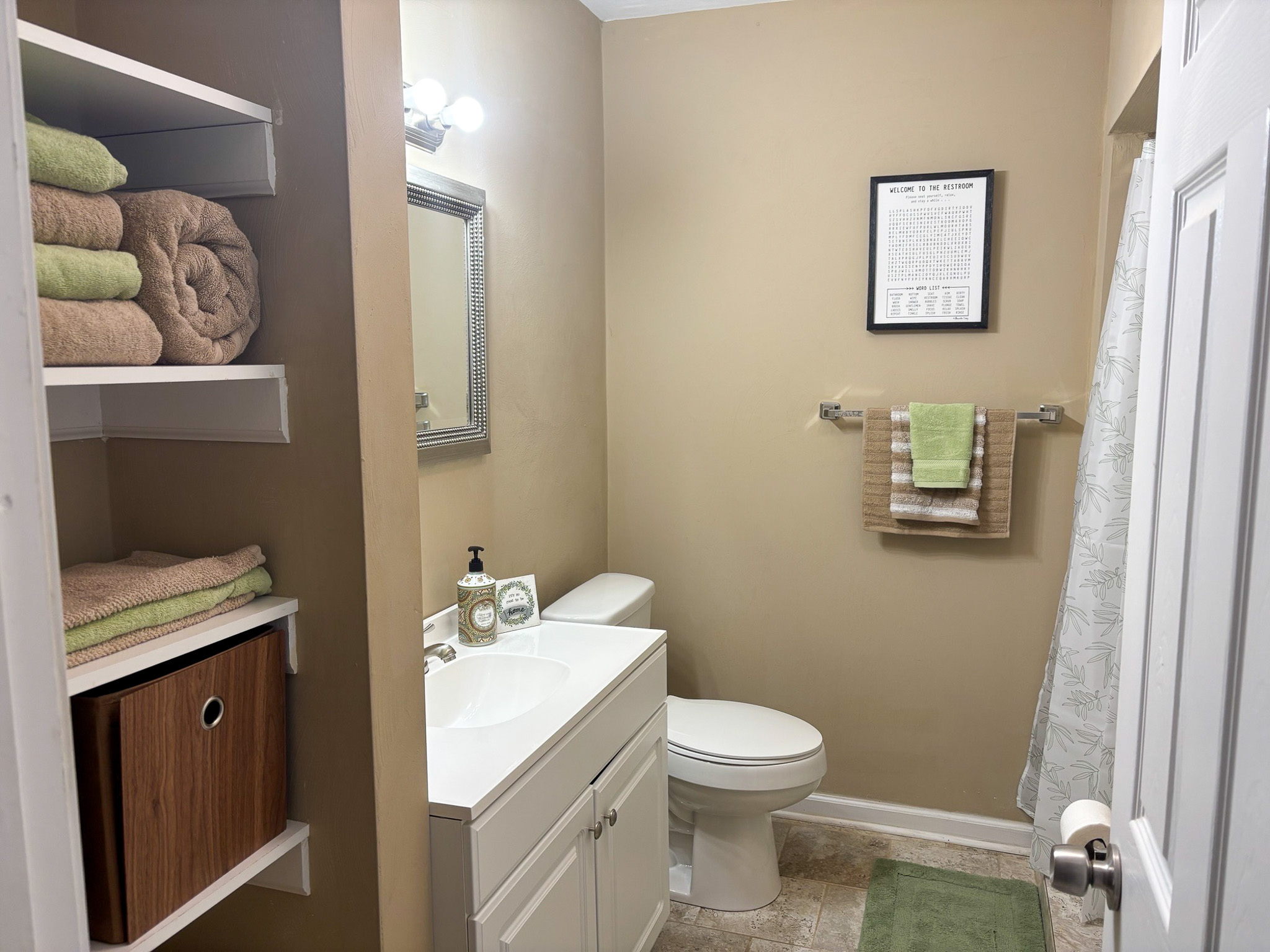 A small bathroom with open shelving holding folded towels, a white vanity, a toilet, and a towel rack on the far wall inside the home of Sponsored Residential Providers Hadassah Hawks and Antonio Hernandez in Salem, Virginia.