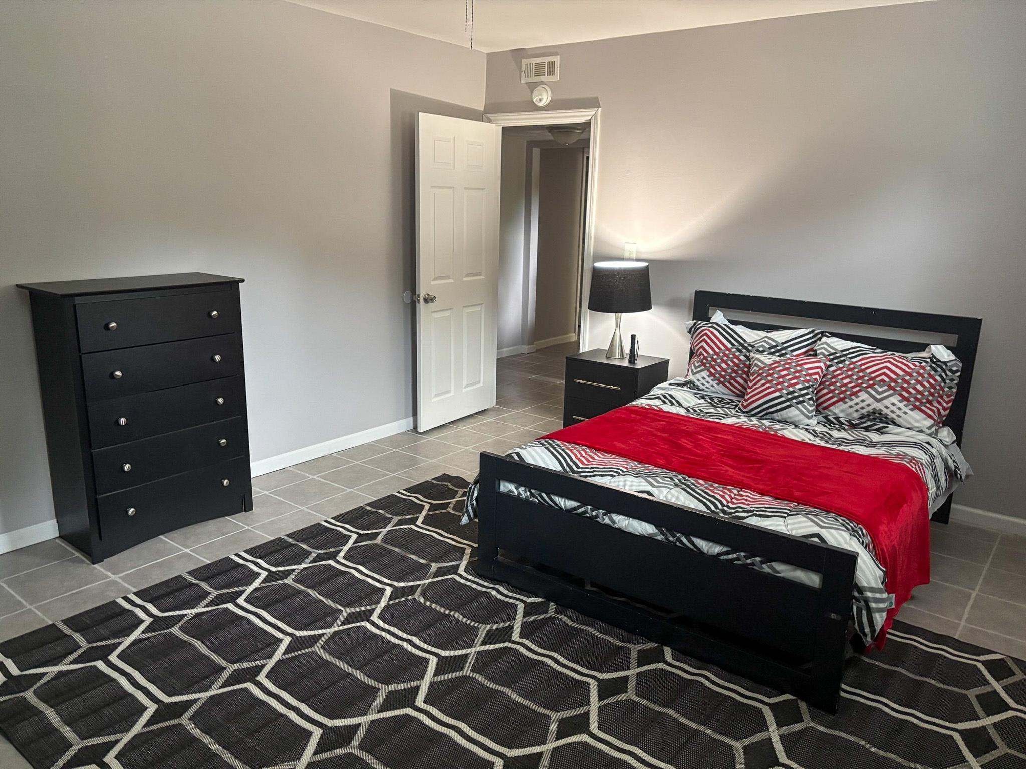 A bedroom with a black bed frame, patterned bedding with a red blanket, a matching dresser, and a large geometric rug inside the home of Sponsored Residential Providers Hadassah Hawks and Antonio Hernandez in Salem, Virginia.