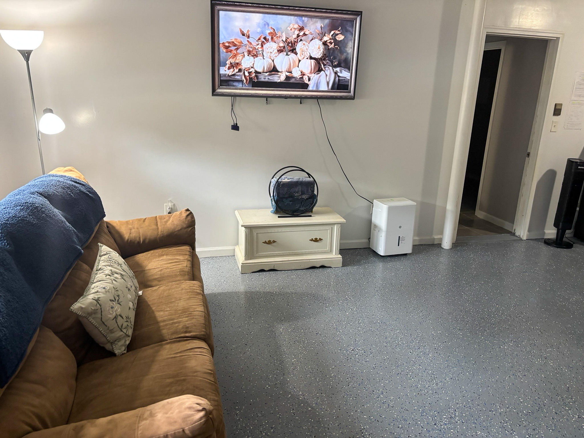 A living room area with a brown couch, a wall‑mounted TV, a small white table with electronics, and a floor lamp inside the home of Sponsored Residential Providers Hadassah Hawks and Antonio Hernandez in Salem, Virginia.