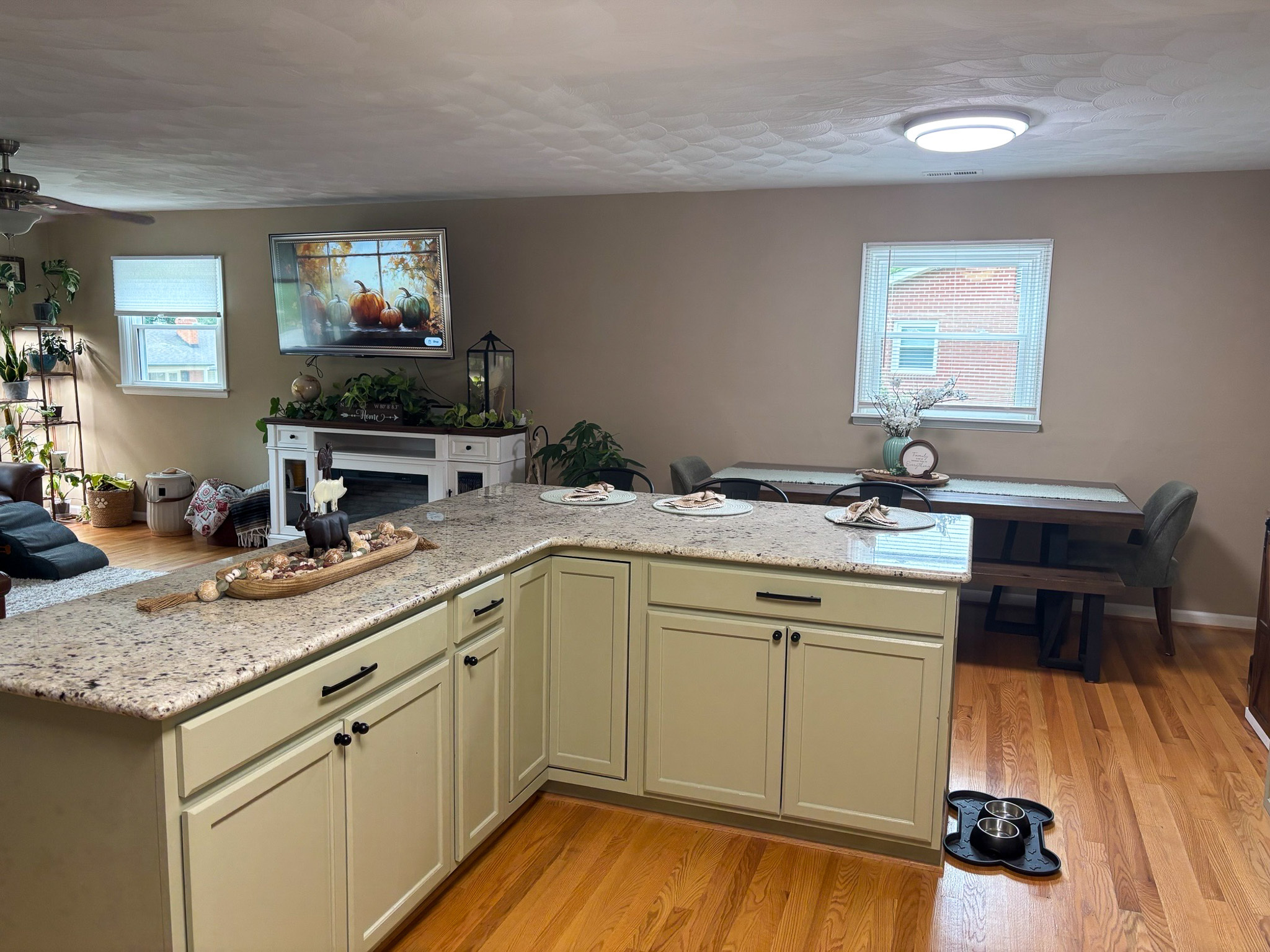 An open kitchen and dining area with a granite peninsula, cream cabinets, a wooden dining table by two windows, and a living space visible in the background inside the home of Sponsored Residential Providers Hadassah Hawks and Antonio Hernandez in Salem, Virginia.