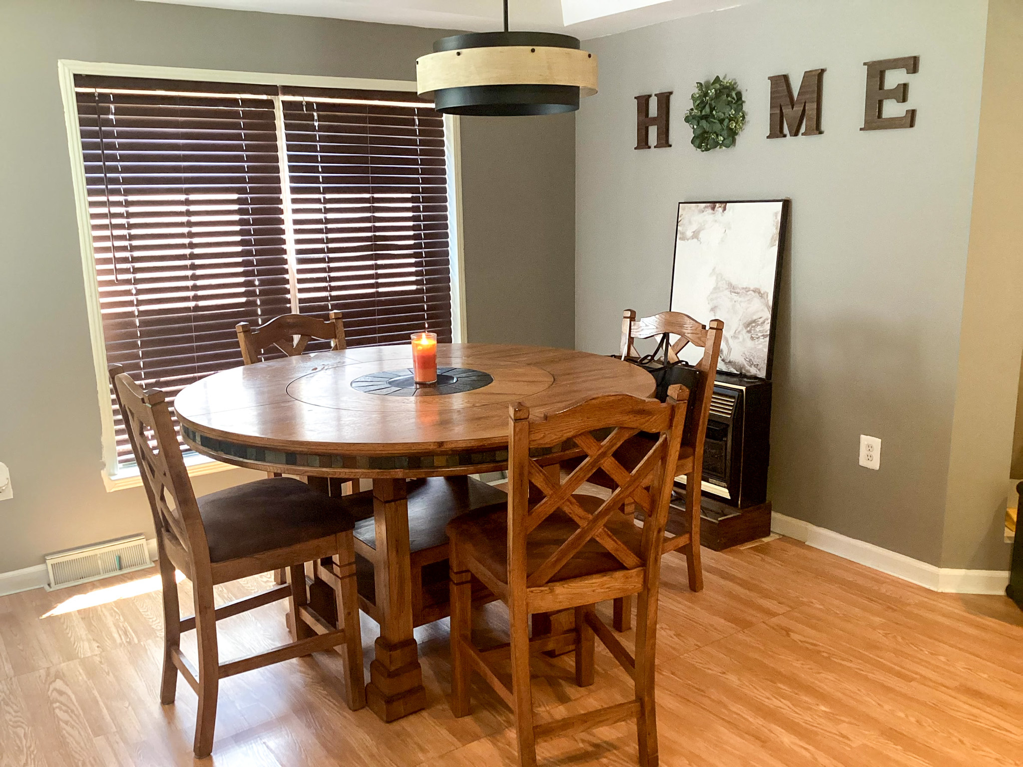 Dining area with a round wooden table, matching chairs, wall art spelling “HOME,” a standing mirror, and window blinds inside the home of Sponsored Residential Provider Kristen Hensley in Madison County, Virginia.