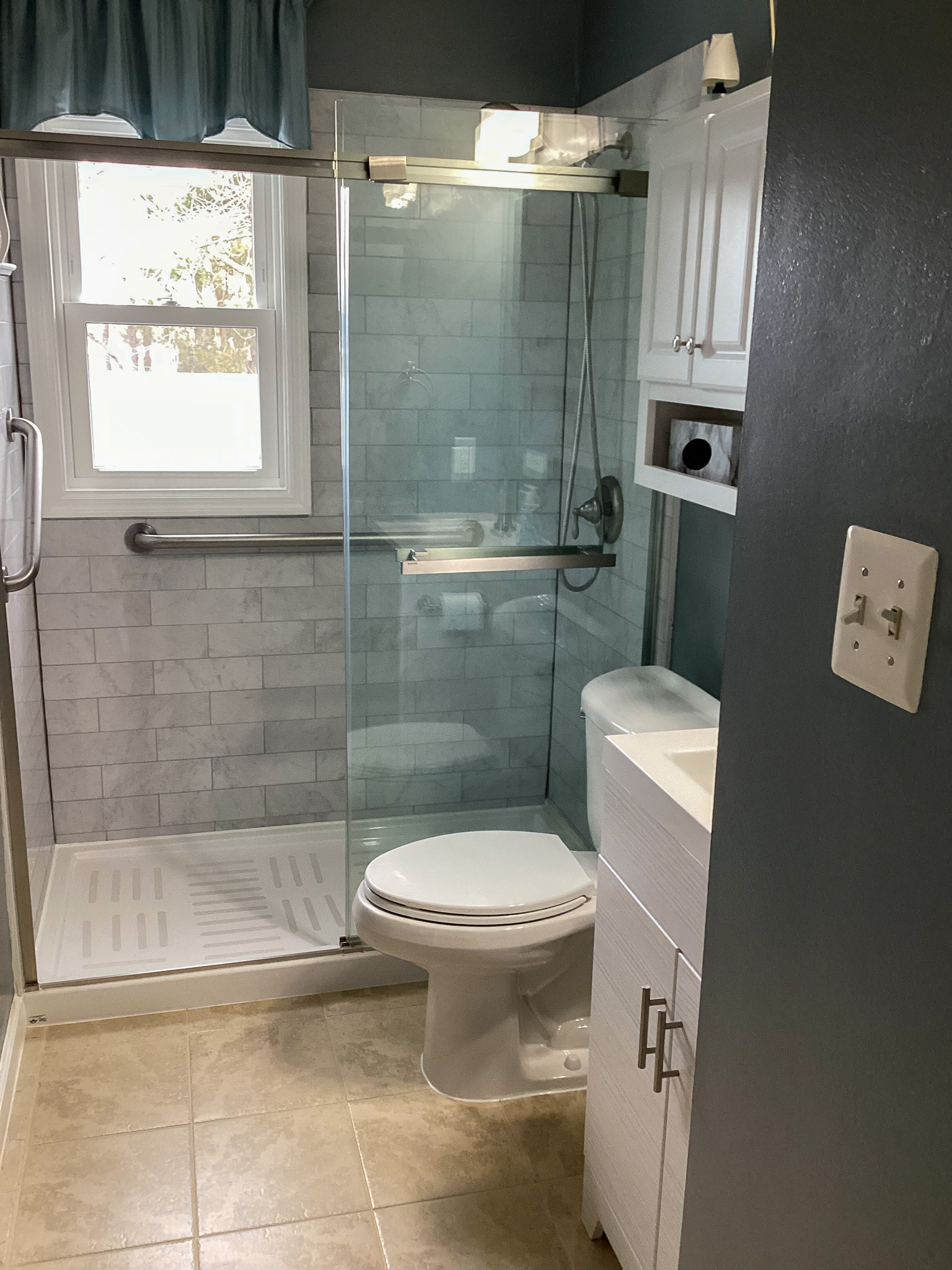 A bathroom with a glass‑enclosed walk‑in shower, a toilet, and a white vanity beneath a cabinet, with a window above the shower letting in natural light, inside the home of Sponsored Residential Providers Teresa and Billy Herran in Woodbridge, Virginia.