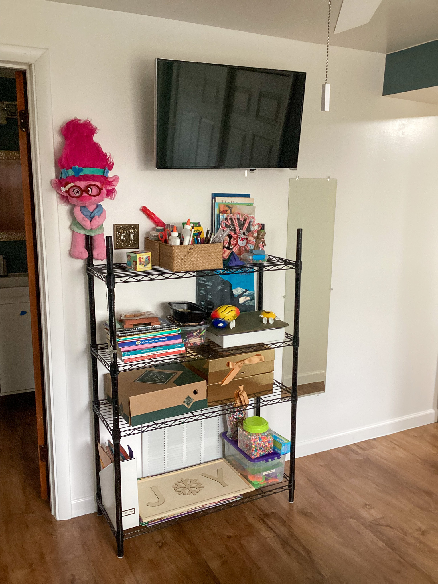A metal shelving unit filled with art supplies, books, and storage boxes stands against a white wall, with a TV mounted above it and a mirror to the right, inside the home of Sponsored Residential Providers Teresa and Billy Herran in Woodbridge, Virginia.